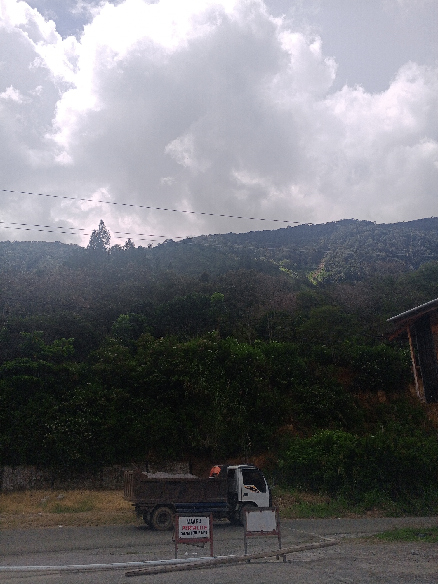 The image shows a scene in the city of Gegarang, Indonesia. In the foreground, there is a dark-colored dump truck parked on the side of a road. The truck is loaded with gravel and appears to be empty of its usual cargo. A person, partially visible in an orange vest, is standing near the back of the truck, possibly overseeing its operation or resting. In front of the truck, a white sign with red text is placed on a stand. The text on the sign reads "MAAF, PERTALITE DALAM PENGIRIMAN." This translates to "SORRY, PERTALITE IS IN TRANSIT."
The background of the image is dominated by a lush, green mountainside covered in dense vegetation. Several power lines are strung across the frame, running horizontally in front of the mountain. The sky above is overcast with dramatic, grey clouds, suggesting potential for rain or a cloudy day. A portion of a wooden building with a dark roof is visible on the far right edge of the image. The overall impression is of a rural or semi-rural environment, possibly a construction site or a transport hub, located in a mountainous region of Indonesia.