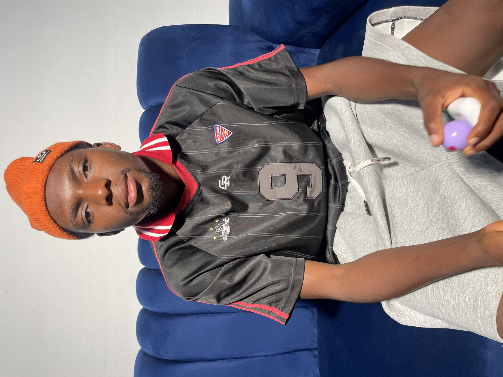 A young man is reclining on a blue couch. He is wearing an orange beanie, a black jersey with red trim and the number 6 on it, and gray sweatpants. He is holding a small, white object with a purple sphere at the end. The background is a white wall. The location is speculated to be Jos, Nigeria, but there is no visual confirmation of this.