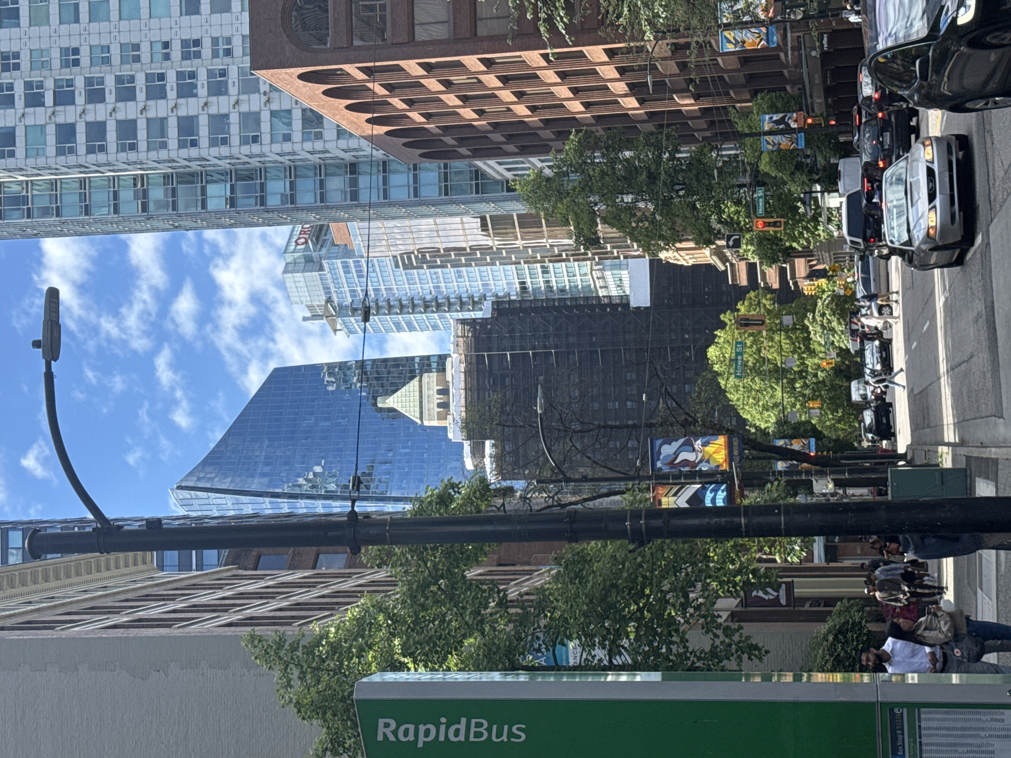 The image shows a street scene in Vancouver, Canada, likely in the downtown core. It is a sunny day with some clouds in the sky. On the left side of the image, a green "RapidBus" sign is visible, indicating a transit stop. Several people are standing on the sidewalk near the bus stop.
The street is lined with tall buildings of various architectural styles. To the left, a light-colored building with dark windows is prominent, with intricate detailing along its top. Behind it, a modern, angled glass skyscraper reflects the sky. In the center background, a darker building under construction, with scaffolding and black netting, is visible, topped with a light-colored dome structure. To the right, a brick building with arched windows stands out. Further down the street, more modern skyscrapers can be seen.
Vehicles are present on the street, including several cars and what appears to be a van. Traffic lights are visible, and one shows a red light for a street labeled "Howe St." Colorful banners with abstract designs are hanging from lampposts along the street. Lush green trees are interspersed among the buildings, providing shade. The overall impression is a bustling urban environment with a mix of old and new architecture.