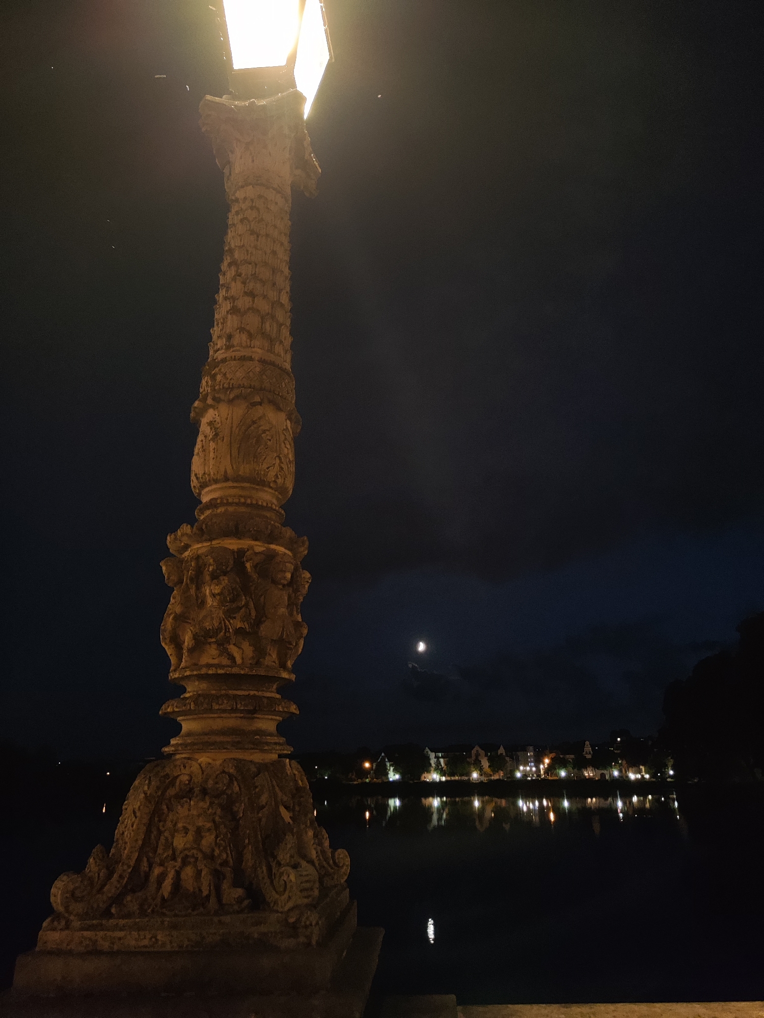 A lamppost with intricate carvings stands in the foreground, its bright light illuminating the dark night sky. In the background, across a body of water, a row of buildings with illuminated windows is visible, their reflections shimmering on the water's surface. The moon is visible in the cloudy sky. The scene appears to be a promenade or waterfront area in Schwerin, Germany, likely near the historic town center.