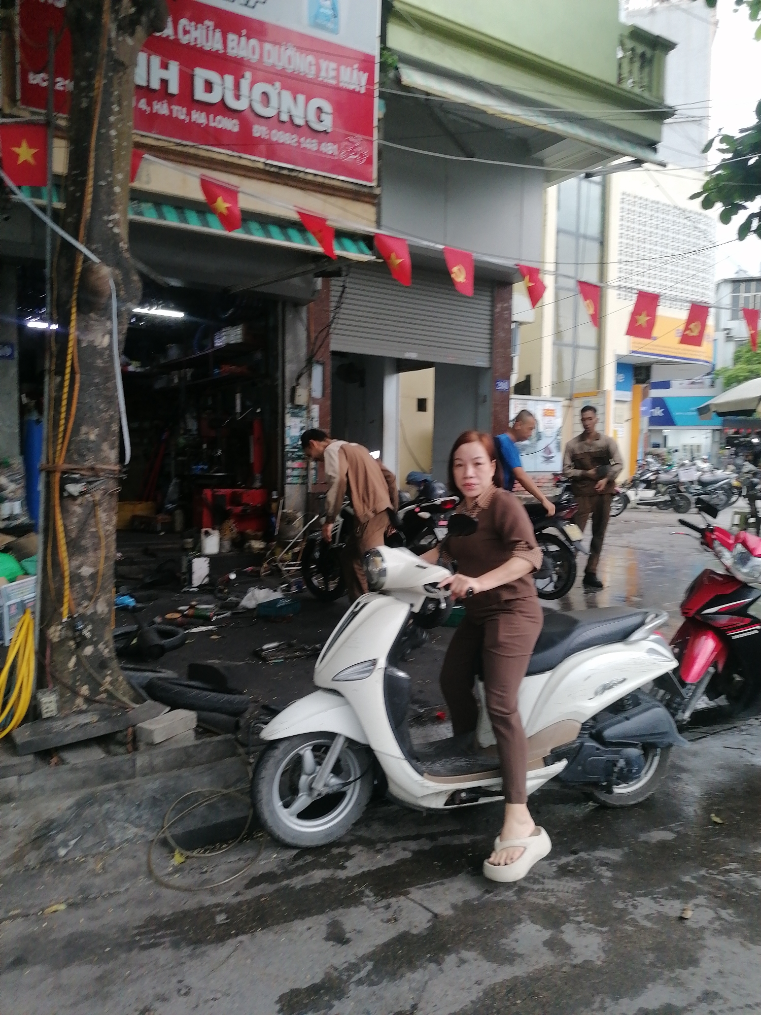 The image captures a street scene in Ha Tu Ward, Vietnam. A woman in a brown jumpsuit and white platform sandals is seated on a white scooter. Behind her, a man in a blue shirt stands near another scooter. Further back, a man in a brown uniform walks on the sidewalk near parked motorcycles. An open garage or repair shop is visible on the left, with a man in a brown shirt working on a motorcycle within. The street appears wet, and a large tree stands beside the garage. A red sign above the garage reads "A CHỮA BẢO DƯỠNG XE MÁY IH DƯƠNG" in Vietnamese. Red flags with yellow stars are strung along the buildings and street.