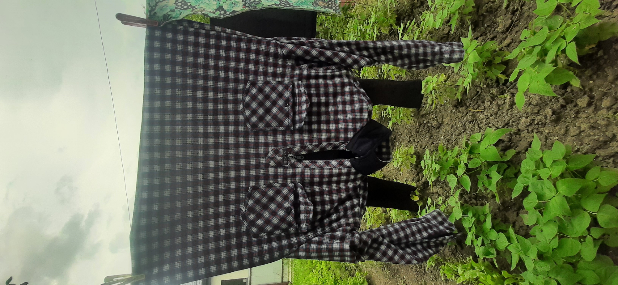 A plaid shirt is hanging on a clothesline outdoors, presumably in Yessentukskaya, Russia, against a backdrop of green foliage and a cloudy sky. The shirt is fastened with a zipper, and a black face mask is visible hanging from it. Two dark-colored pant legs are visible hanging from the bottom of the shirt, suggesting it is being worn or hung in a way that mimics a person. The shirt is predominantly maroon, white, and black plaid. Two chest pockets are visible on the shirt. The surrounding environment consists of tilled soil and numerous small green plants. The image is taken from a slightly low angle.