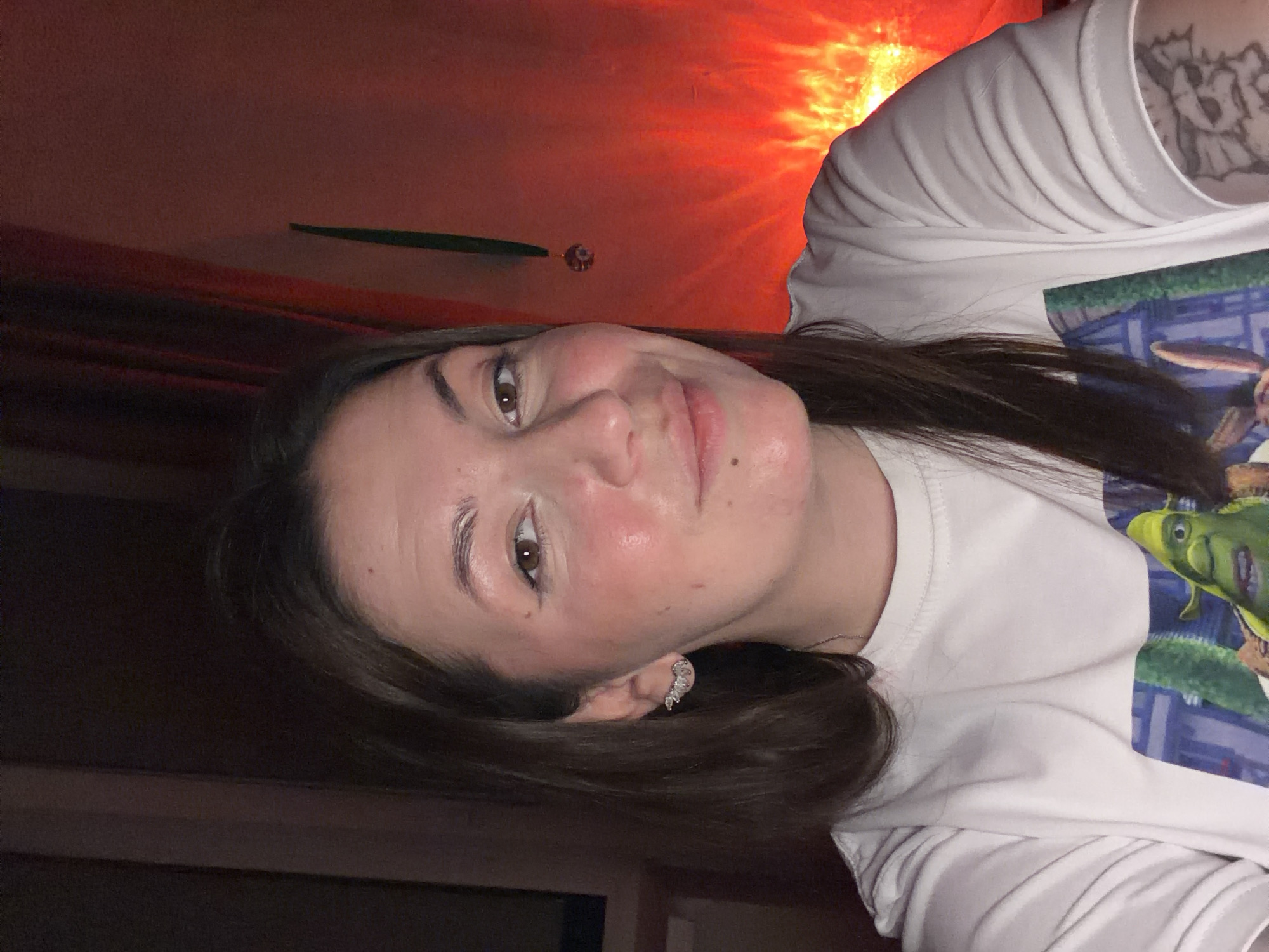 A woman with dark hair is centered in the frame, angled slightly to the right. She is wearing a white t-shirt, partially visible. An earring is visible on her left ear. Behind her is a reddish-orange light source, possibly a lamp. In the background, a glimpse of a green object is visible above her head, along with a crystal-like ornament. There is no indication of a specific location in the city of Chaikovsky, Russia.