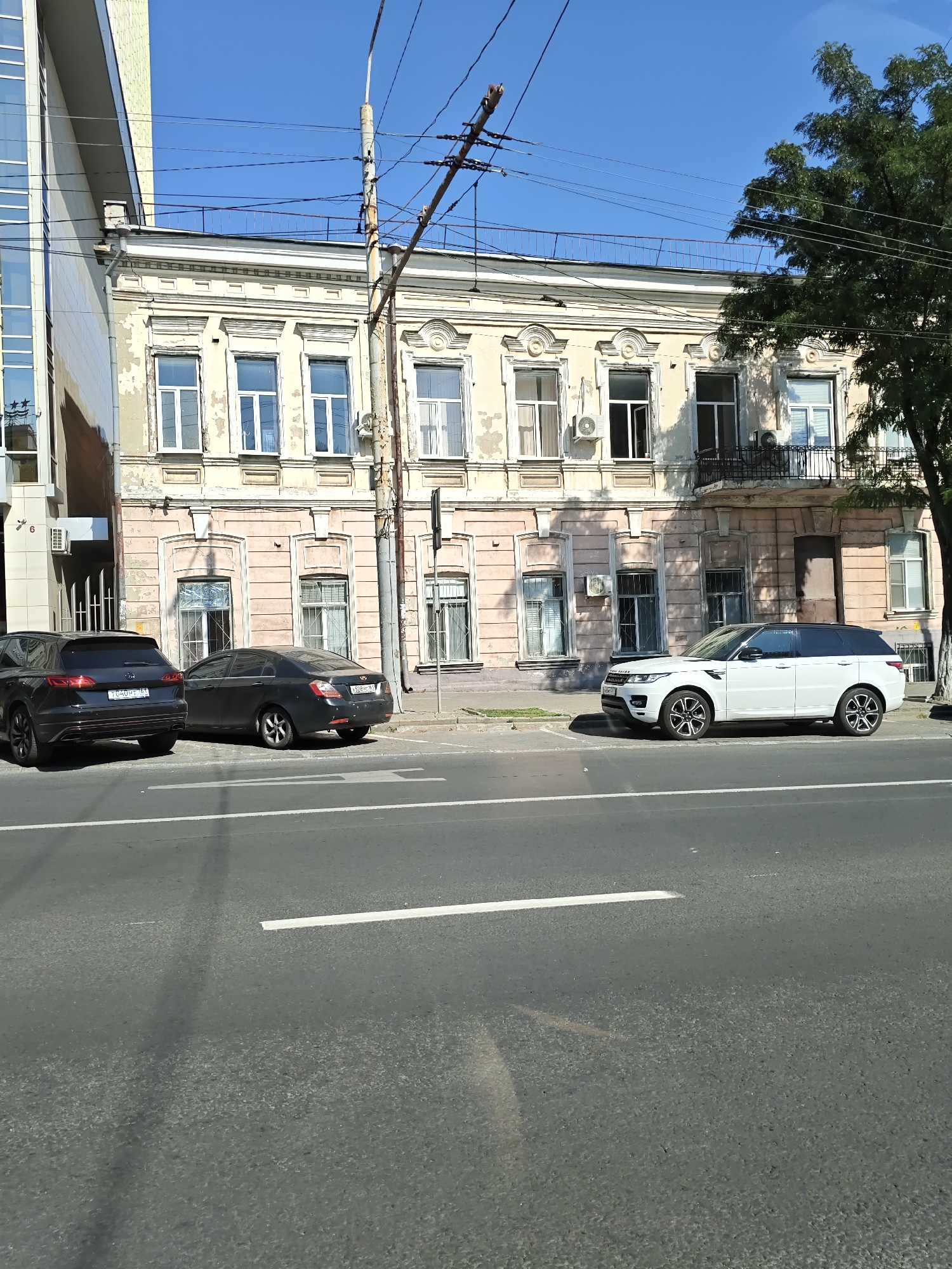 A street scene in Rostov-on-Don, Russia, on a sunny day, features a multi-story building with a pale yellow facade and a pink lower level, characterized by ornate architectural details, multiple windows, and air conditioning units. To the left, a modern glass and metal building stands adjacent. In front of the older building, three cars are parked: a dark SUV on the left, a dark sedan in the middle, and a white SUV on the right. The street in the foreground is paved, with white lane markings. Overhead, power lines and a utility pole are visible against a clear blue sky. A tree with dark green foliage is on the right side of the frame.
