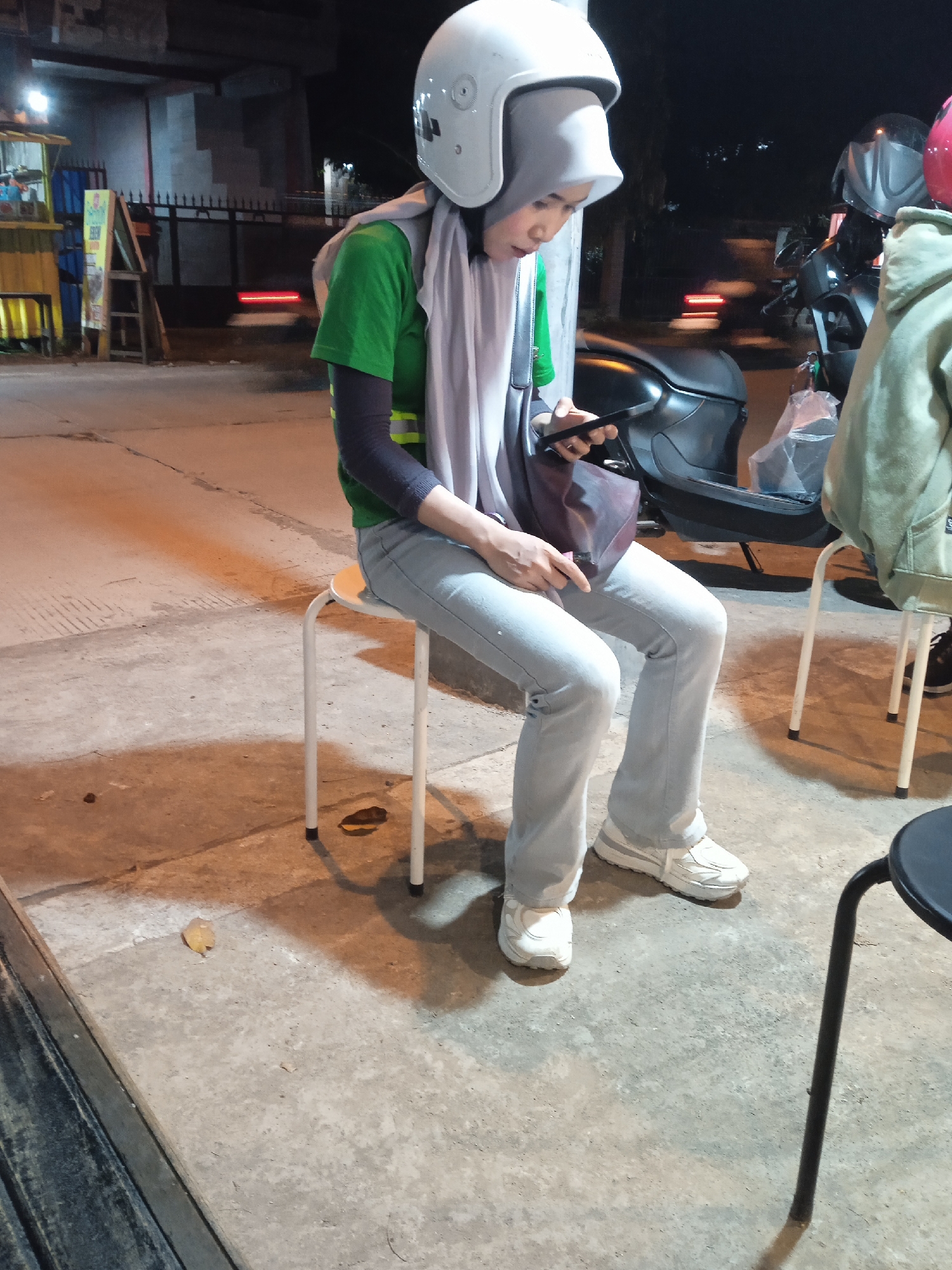 This nighttime image captures a candid moment on a street in Sekopek Wetan, Indonesia. The central figure is a person seated on a white stool, intently looking down at a smartphone held in their left hand. They are wearing a white full-face motorcycle helmet, a light gray hijab, a green t-shirt layered over long black sleeves, light blue jeans, and white sneakers. A dark red/brown shoulder bag is positioned on their lap.
To the right, another person is partially visible, wearing a khaki or light green hooded jacket and seated on a black stool. Behind them, a black scooter or motorcycle is parked, with a plastic bag hanging from its handlebar. Another motorcycle, partially visible, has a red helmet resting on it.
The background is dark, illuminated by artificial streetlights and possibly lights from the vendor stall. On the far left, a small yellow vendor cart or stall with shelves and some indistinct signs is present. Next to it, a wooden easel-like stand displays faded, unreadable text and an image, likely an advertisement or menu. Further back, concrete pillars or supports for an overpass or bridge are visible, along with a dark fence. A blurred red light streak in the middle ground suggests a moving vehicle on the road. The ground is a concrete pavement with some cracks and reflects the ambient light.
The scene depicts a casual, urban street environment at night, possibly a waiting area or a spot near a local eatery, with the person focused on their device. No specific text is clearly legible in the image.