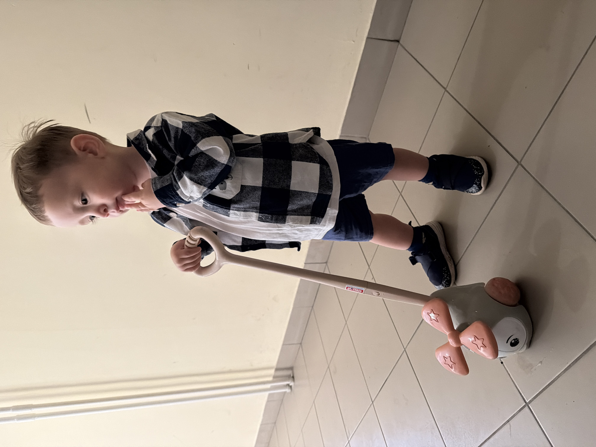 A young boy, approximately 2-3 years old, stands indoors on a tiled floor, holding a toy airplane push toy with a pastel pink propeller and wheels. The boy is wearing a white t-shirt under a navy and white checkered shirt, navy blue shorts, and dark blue sneakers with white soles. He is looking to his left with his right index finger touching his chin. The background consists of a light yellow wall with white vertical pipes to the left and a tiled baseboard. The overall scene appears to be within a residential or public building.