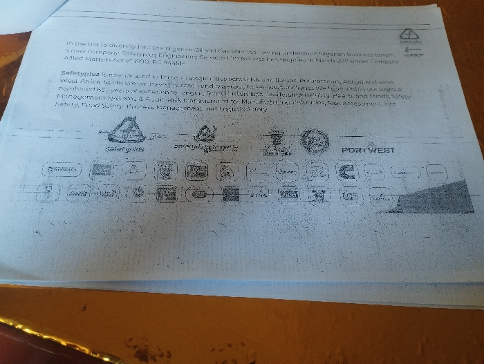 The image is of a document on a blue piece of paper. The document describes the services of Safetyplus Engineering Services Limited, a new company incorporated in March 2011 under Company Allied Matters Act of 1990 (RC 834464). There are two triangular diagrams with the words “safety skills” and “emerging technologies”. The image contains several smaller logos below these, and on the right hand side of the image it reads “Port West”. The paper appears to be on a wooden table.