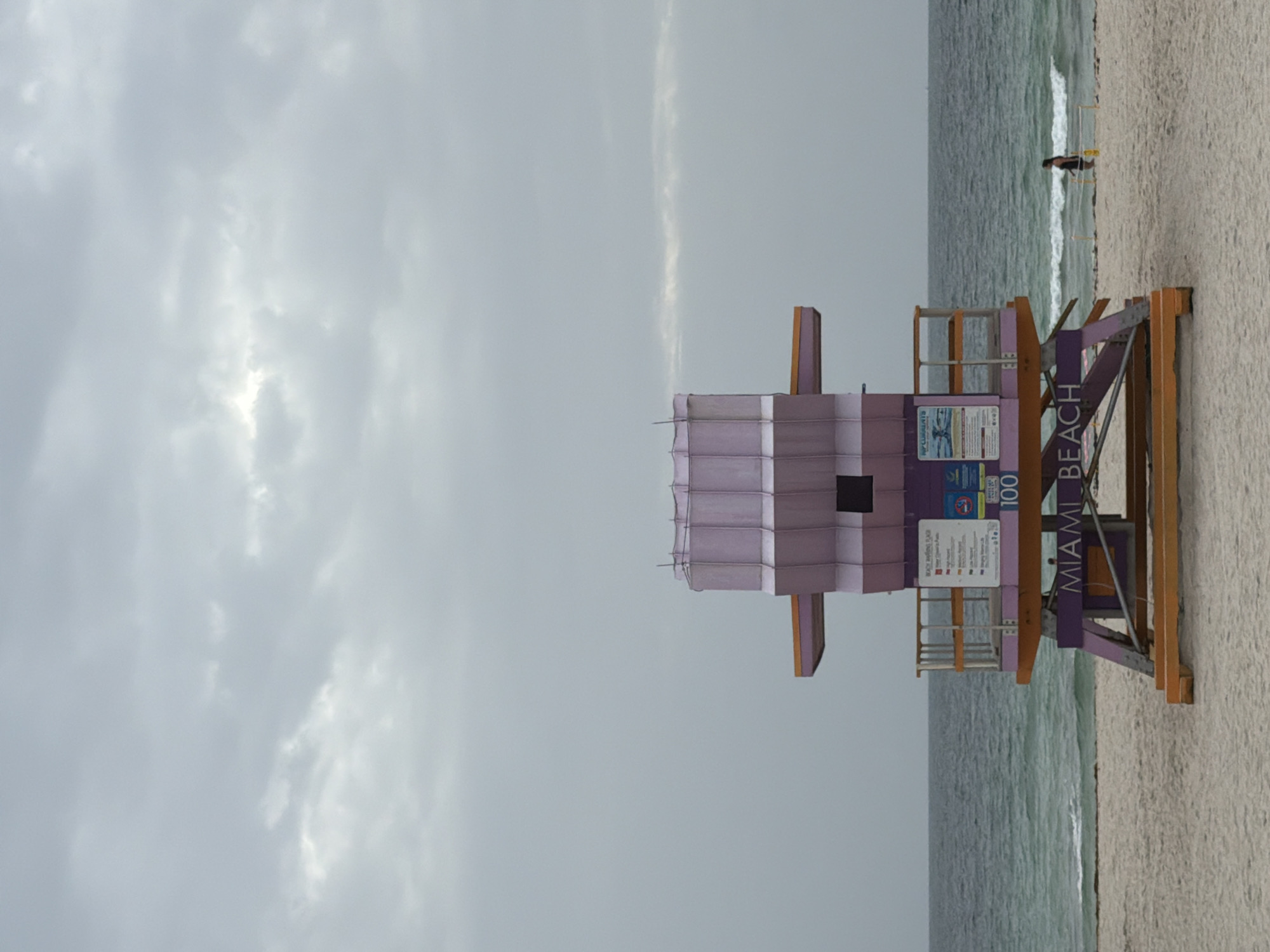 A lifeguard tower stands on a sandy beach in Miami Beach, Florida, under a cloudy sky. The tower is painted purple and orange, with a purple section at the top and a purple base. The word "MIAMI BEACH" is visible in purple letters on the base of the tower. The ocean is to the left of the tower, with waves breaking on the shore. A single person, a woman wearing a black swimsuit, is walking on the beach to the right of the tower, facing away from the camera. The sand is light brown and appears damp in some areas. The sky is overcast with a mix of light and dark gray clouds.