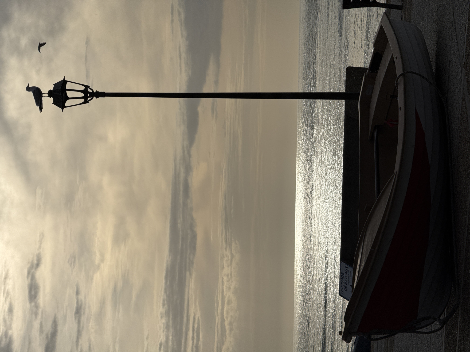 The image presents a coastal scene in Étretat, France, captured during low sun, likely sunrise or sunset, under an overcast sky. The sun's light is diffused by multiple layers of stratocumulus clouds, which display gradients of light grey, beige, and soft orange hues. The sea, occupying the mid-ground, reflects the intense sunlight, creating a shimmering silver and white expanse across its surface.
A tall, dark, ornate lamppost stands prominently in the central mid-ground, extending from near the bottom of the frame almost to the top. A large seagull is perched atop the lamppost's lantern, silhouetted against the bright sky, with its head turned to the right. A second, smaller seagull is captured in flight in the upper-right quadrant, also silhouetted.
In the immediate foreground, a small, clinker-built wooden boat rests on a pebbled or gravelly surface. The boat's hull is largely darkened by backlighting, but a distinct red stripe is visible on its bow above a lighter-toned lower section. A coiled rope is present near the boat's front. Behind the boat, a low, dark concrete wall or barrier extends horizontally. A white sign with black lettering, partially obscured by the boat but legible, displays the text "ECHOUAGE DE BATEAUX," which translates to "BOAT STRANDING/BEACHING." A dark, wooden bench is partially visible on the far right edge of the frame, positioned behind the boat. No human figures are present in the scene.