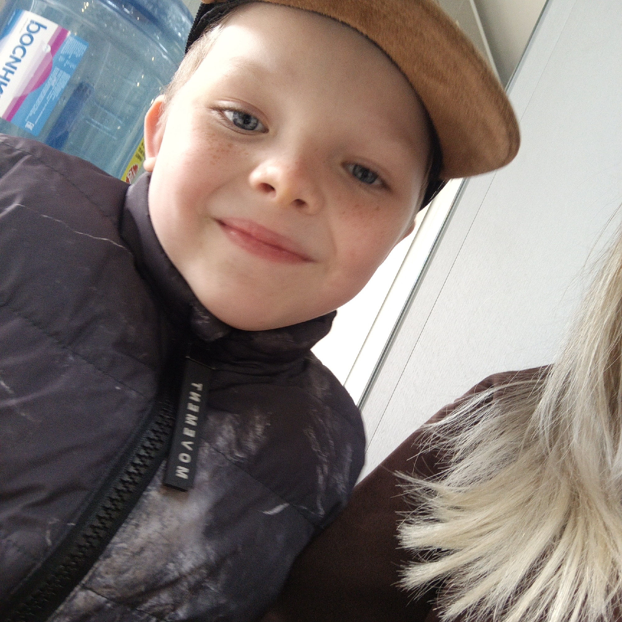 The image shows a close-up shot of a young boy with fair skin, freckles, and blue eyes. He is wearing a brown beret and a black, patterned jacket with a zipper that has a tag reading "MOVEMENT." A portion of a large, blue water cooler is visible in the background, along with a section of a white wall. To the right, there is a blurry section of blonde hair and a dark brown garment or piece of furniture. The specific location and city are not discernable from the image.