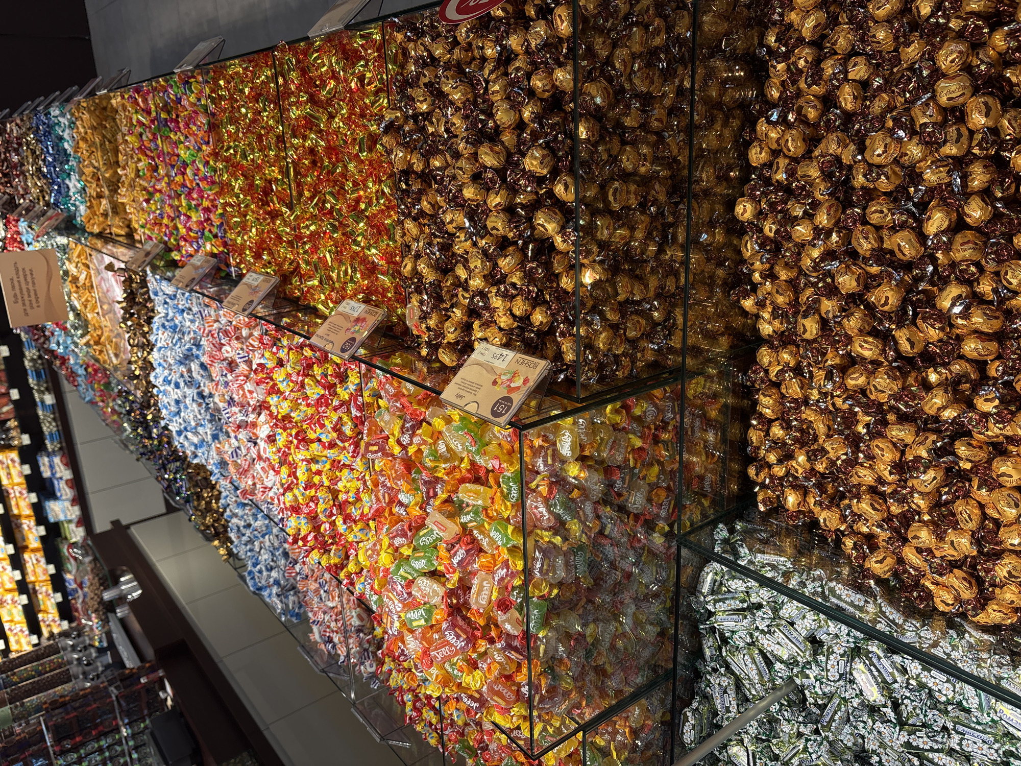 The image shows a candy display in what appears to be a store in Hatne, Ukraine. A variety of candies are displayed in clear containers, likely made of glass or plastic. The candies are individually wrapped and come in different colors and shapes. Some of the candies are wrapped in gold foil. There are price tags on some of the containers. A sign is visible in Ukrainian, instructing customers to separate the candy types into different bags for weighing.