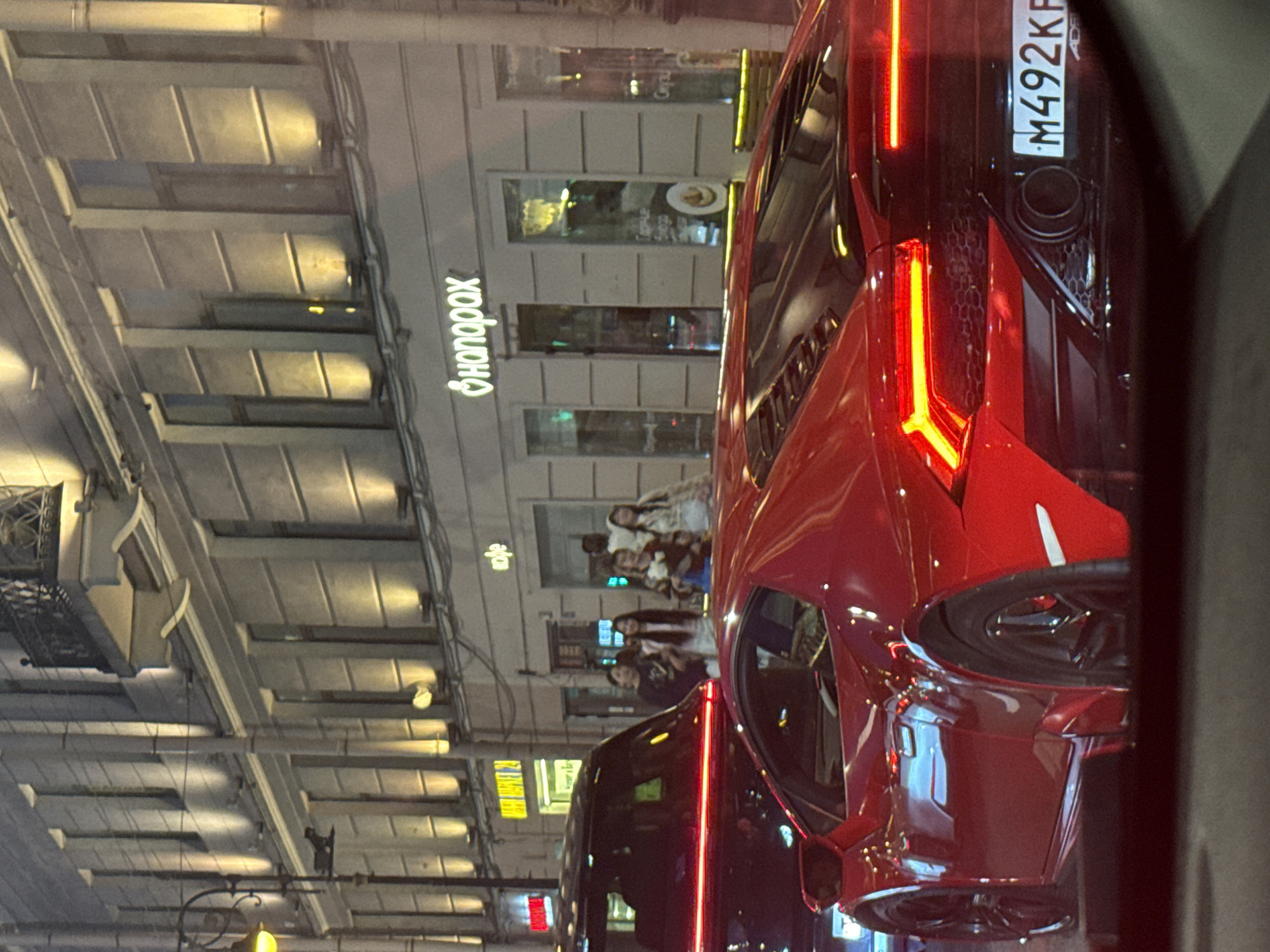 The image depicts a street scene in Saint Petersburg, Russia, at night. In the foreground, a red sports car, likely a Lamborghini Huracán, is parked. Its rear lights are illuminated, and a license plate reading "M492KP" is visible. To the left of the sports car, the back of a black SUV or similar vehicle is seen, with a red light bar across its rear.
Behind the vehicles, a building with multiple illuminated windows and architectural details stands. A neon sign with Cyrillic text, appearing to be "ФОНОРАХ" (Fonorakh), is mounted on the building. Several people, appearing to be young women, are walking on the sidewalk in front of the building, looking towards the camera or engaging with each other. The street is lit by streetlights and the lights from the buildings. The overall impression is a city street at night, with prominent vehicles and urban architecture.
