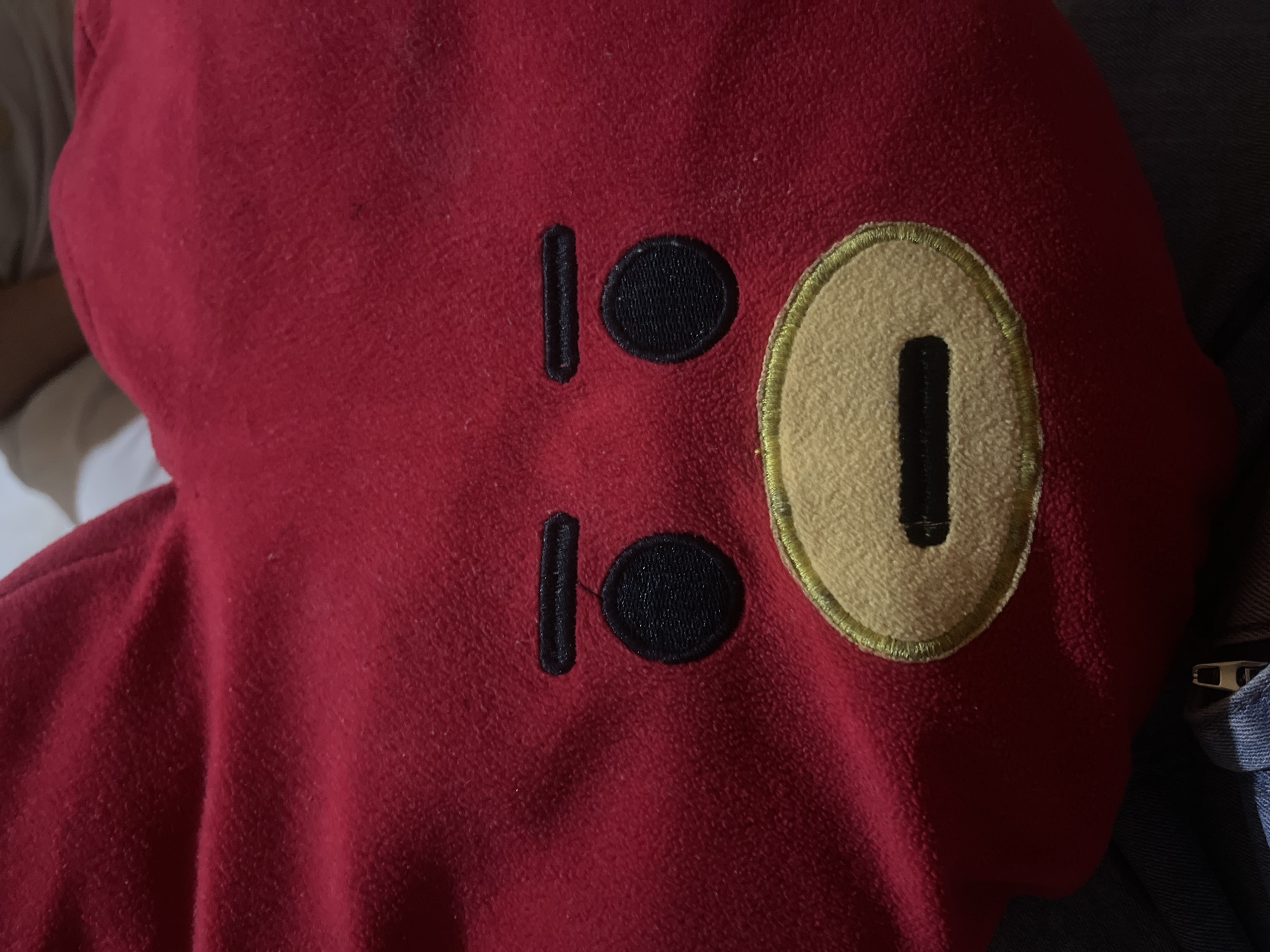 The image shows a close-up of a red garment, possibly a hoodie or jacket. Two black embroidered shapes, resembling eyes, are positioned vertically on the left side of the garment. To the right of these, a larger, oval-shaped embroidery in a tan color with a yellow embroidered outline contains a single black embroidered shape, resembling a mouth or a nose. The texture of the red fabric appears soft and slightly fuzzy. The garment is resting on a surface, and a portion of blue denim fabric is visible on the right edge of the frame, with a zipper detail. The lighting suggests an indoor setting, and the overall impression is that of a close-up detail of clothing. There is no visible text in the image.