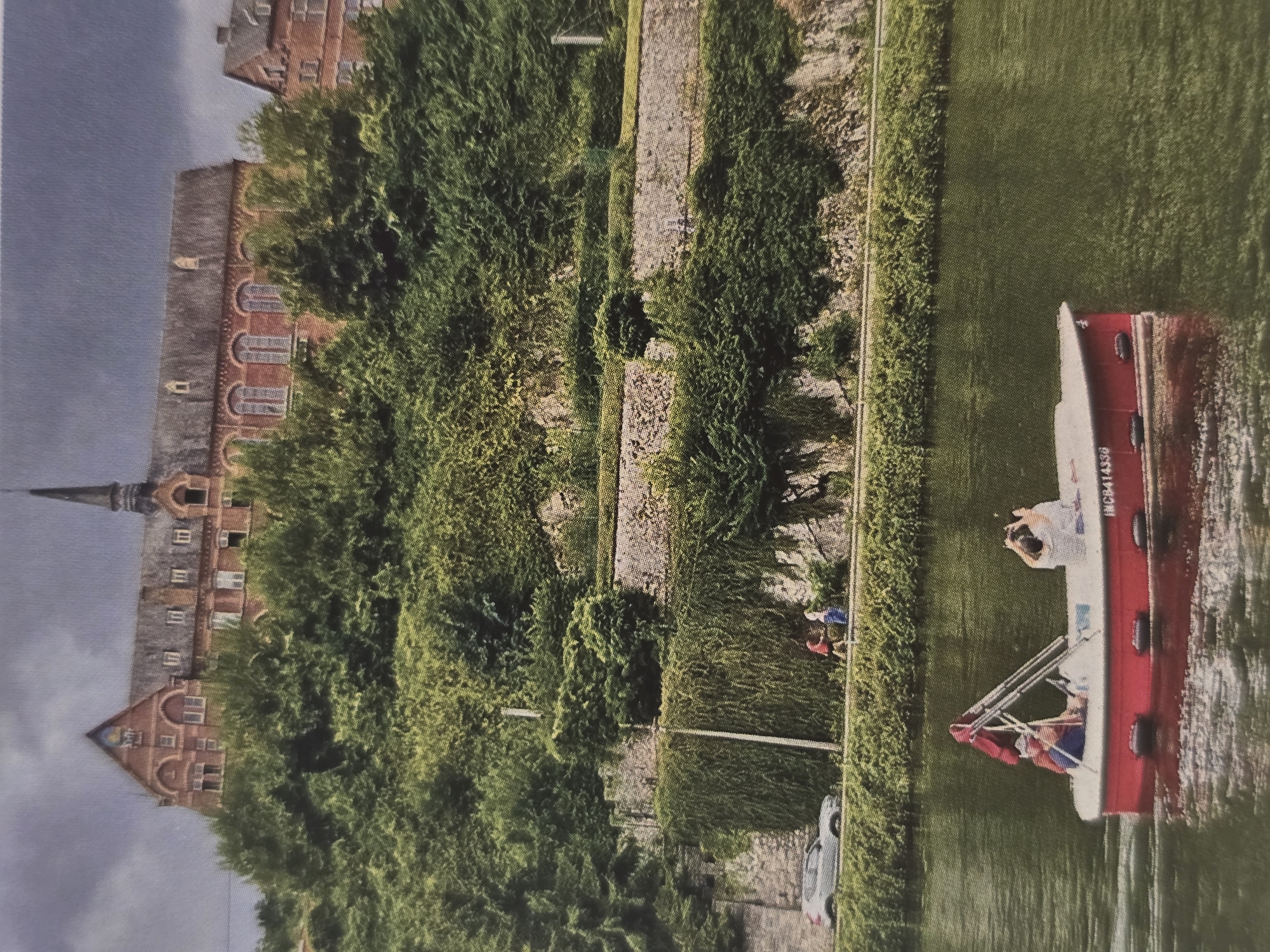The image depicts a tranquil daytime scene along a waterway, likely a canal or river, set in Sofia, Bulgaria. The sky is overcast, casting a soft, even light over the landscape.
On the right side of the frame, a red and white small motorboat or leisure craft cruises on the water. A person in a white top stands at the stern, looking away from the camera, possibly observing the scenery or taking a photo. Another individual is partially visible towards the bow. The boat bears the registration number "NCB4143B" on its side.
The waterway is bordered by a green, heavily vegetated bank and a stone or paved path. Along this path, in the middle-ground, two people are walking; one wears a red top and blue pants, and the other a blue top. A light-colored car is parked on the path, partially obscured by bushes. Several utility poles are also visible along the path.
On the far left, behind a dense line of trees and bushes that cover a hillside, stands a large, multi-story brick building. Its architecture appears old, featuring numerous windows, some arched, and a distinctive dark spire or turret. The building suggests a historical structure, such as a castle, old factory, or institutional building. The overall scene conveys a sense of leisure and natural beauty combined with historical architecture.