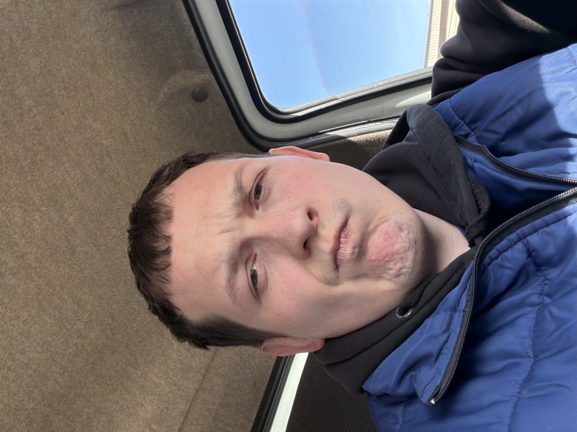 A person is pictured from a low angle, possibly inside a vehicle. A sunroof is visible in the upper portion of the image, allowing a view of a clear blue sky. The vehicle's interior includes a tan-colored fabric covering the ceiling. The individual is wearing a blue puffer jacket over a black hooded sweatshirt. The location in the city of Chaikovsky, Russia is not able to be determined.