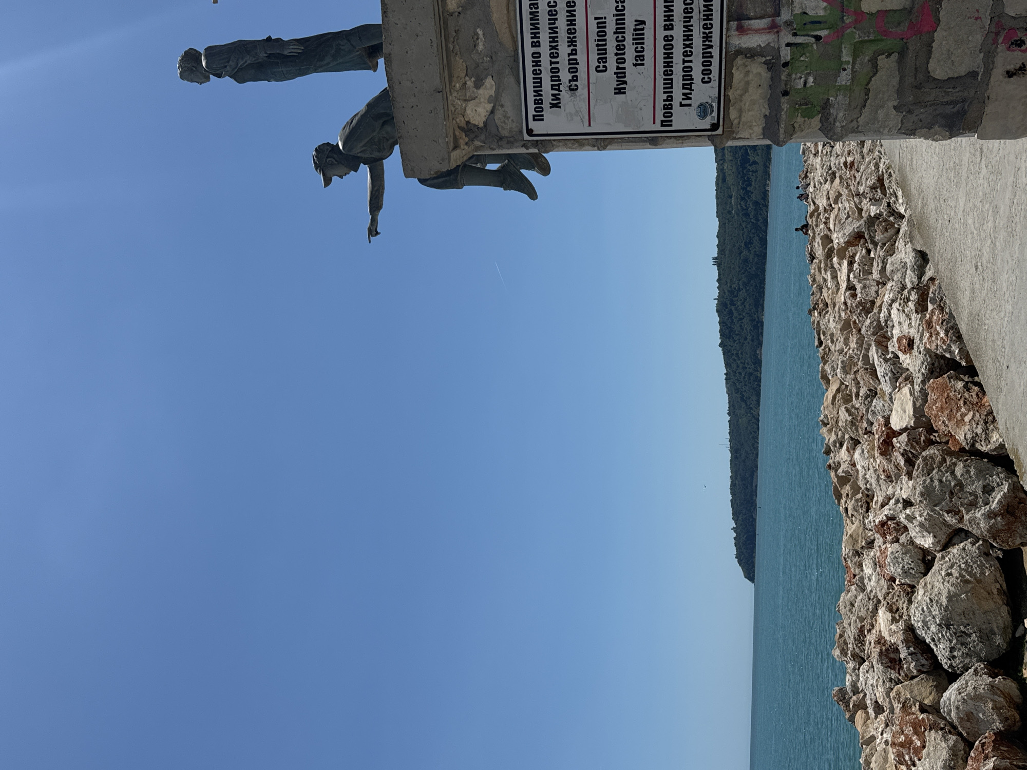 In the city of Unknown, Unknown, this image captures a serene outdoor scene. A clear blue sky serves as the backdrop for a statue of two figures, one seated and pointing towards the horizon, the other standing beside them. Both are rendered in a classic style. Below the statue, a concrete structure displays a multilingual sign warning of a "Hydrotechnical facility" with caution. The foreground is dominated by a rocky embankment leading to the turquoise sea. The water stretches to meet a distant, tree-covered coastline under a bright, sunny sky. A few individuals are visible in the distance, seated on the rocks by the water, enjoying the view. The image is composed from a slightly low angle, emphasizing the height of the statue and the expanse of the sky.