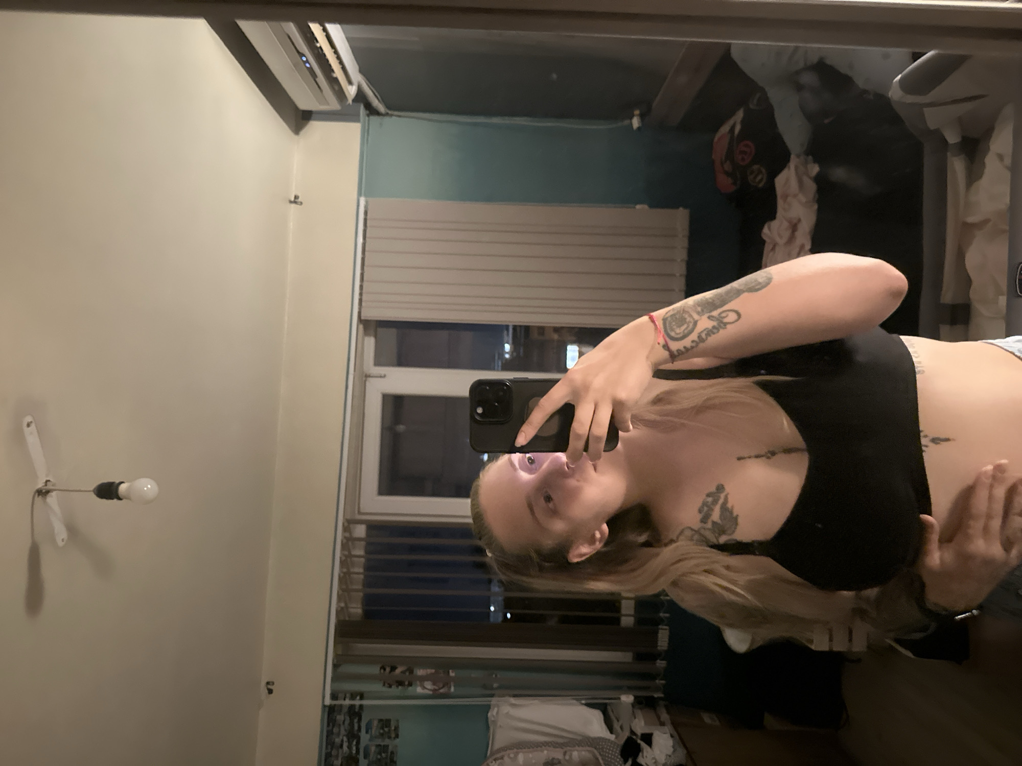 A young woman with blonde hair pulled back in a ponytail stands in a room, taking a selfie in a large mirror. She is wearing a black sports bra and has tattoos on her chest, stomach, and left arm. She is holding a black smartphone in her right hand. The room appears to be a bedroom or living area, with a bed visible in the background. The lighting in the room is dim, with a bare lightbulb hanging from the ceiling casting a shadow. An air conditioning unit is mounted on the wall above a window with vertical blinds. There are several items of clothing and bedding scattered around the room. The overall impression is of a casual, indoor setting. The image does not contain any specific details that would allow for identification of the location within Plovdiv, Bulgaria.
