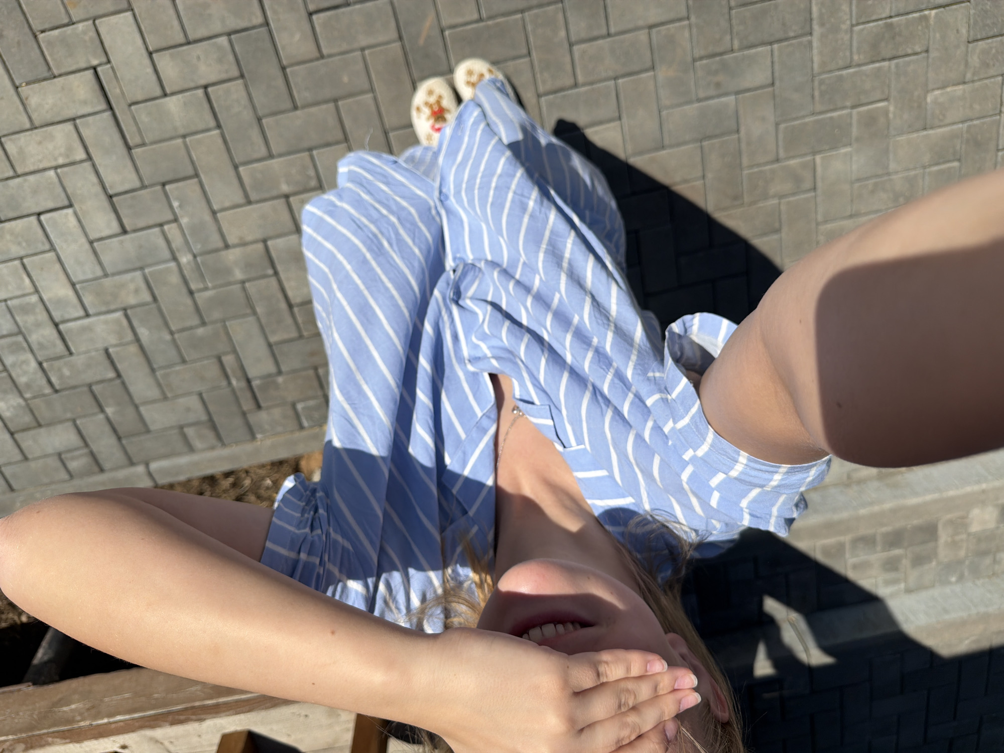 A woman is taking a selfie outdoors in what appears to be Kamennoye, Russia. The image is taken from a high angle, looking down at the woman who is covering part of her face with her hand. She is wearing a blue and white striped dress. Her feet are visible, clad in white slippers. The background consists of a paved area with interlocking gray bricks laid in a herringbone pattern, and shadows are cast by a nearby object.