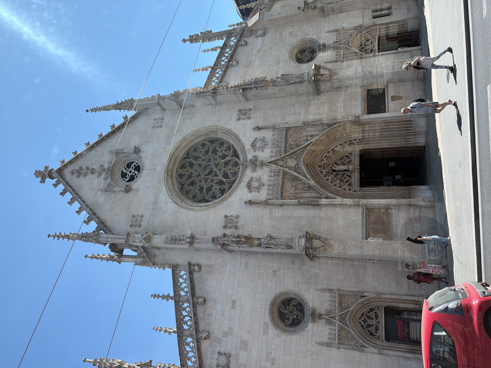 The image shows a street view of the exterior of a large, ornate Gothic-style church with a light-colored stone facade. The church features intricate architectural details, including spires, arched doorways, rose windows, and statues. A bright blue sky is visible above.
In the foreground, a red car is parked on the left side of the street. Several people are walking on the sidewalk in front of the church. On the right side of the image, two people are walking away from the camera, and further left, three people are standing and looking towards the church. A fourth person is walking away from the camera on the far right.
The church in the image is the Cathédrale Saint-Jean-Baptiste de Lyon, located in Lyon, France. The street in front of the cathedral is a public thoroughfare, indicated by the presence of cars and pedestrians. The sunny weather suggests a daytime scene.