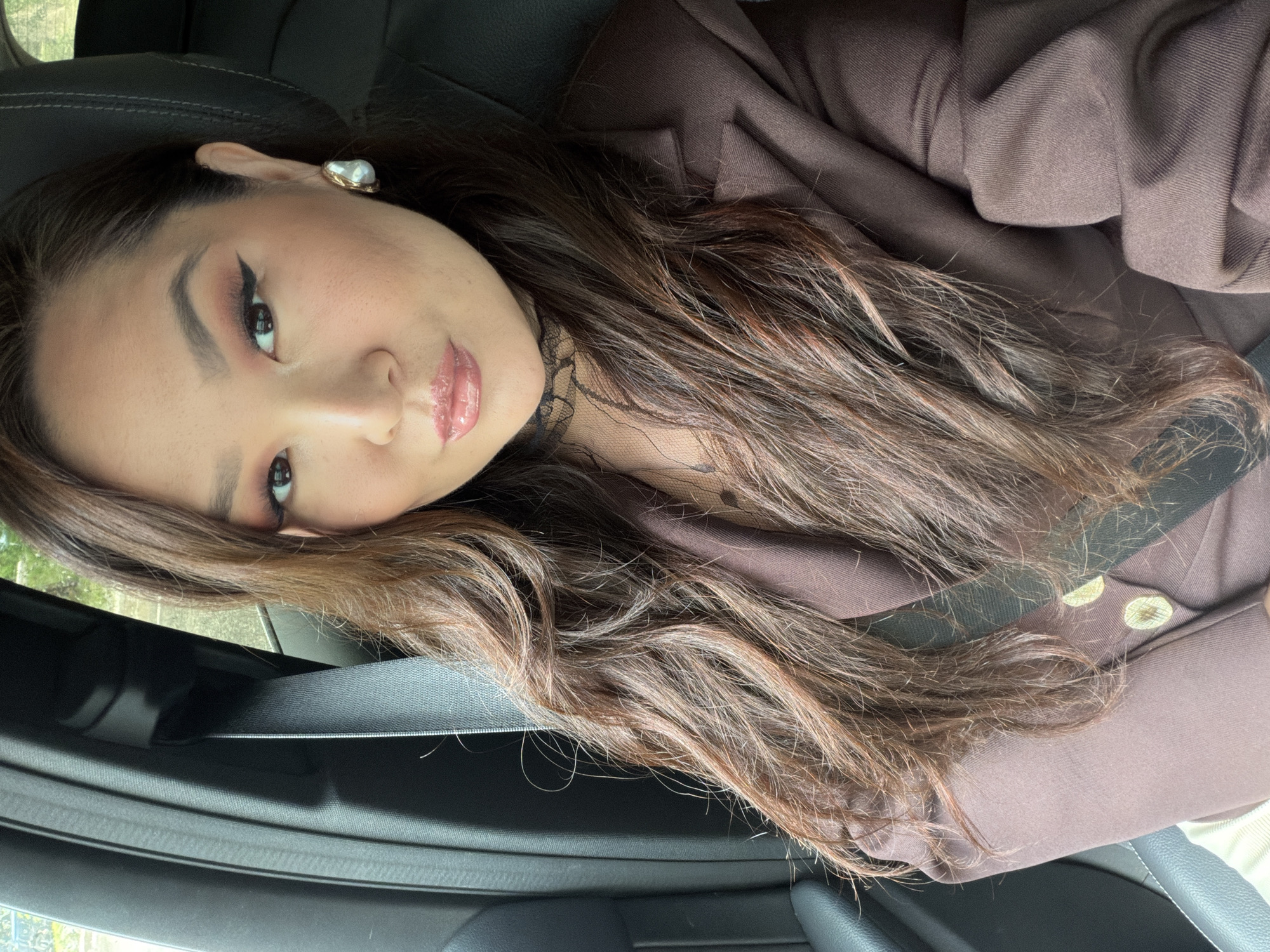 The image presents a close-up, oblique portrait of a young woman seated inside a car, likely in Chaikovsky, Russia, during daytime. Her head is tilted, and she looks towards the camera with a calm, slightly smiling, and posed expression. She has long, wavy brown hair, which appears to have subtle reddish undertones or highlights, flowing over her left shoulder and chest.
The woman is wearing a dark brown blazer or jacket over a black, possibly lace, top, visible at her neckline. A black seatbelt crosses diagonally over her chest. Her makeup is notable, featuring sharp winged eyeliner, coordinated eyeshadow in warm, rosy hues, and glossy, light pink lips. A single, distinct earring is visible on her left ear, comprising an irregularly shaped white pearl set in a gold-toned metal.
The immediate setting is a car interior, with dark upholstered seats and interior trim visible. Through the car window on the left, blurred green foliage suggests an outdoor environment and natural daylight. There is no visible text in the image. The overall scene depicts a personal moment or selfie taken within a vehicle.