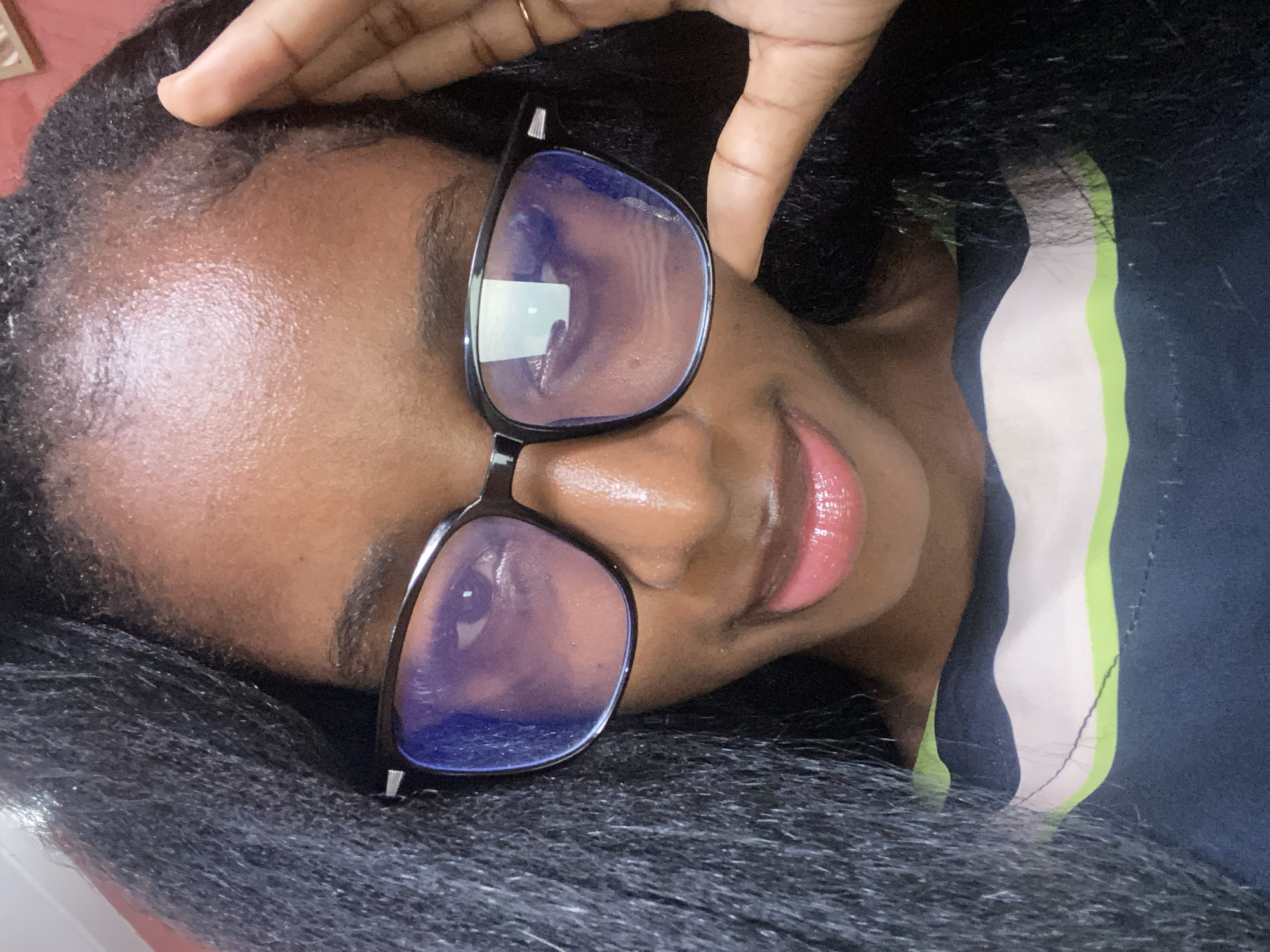 The image is a close-up, waist-up selfie of a young Black woman wearing black-rimmed glasses and a dark blue shirt with horizontal white and lime green stripes. Her long, dark hair frames her face, and her right hand is raised to her temple, with a gold ring on her ring finger. She has pink lipstick on and appears to be looking directly at the camera. The lighting suggests an indoor setting, likely in Jos, Nigeria. The background is blurred and appears to be a solid color, possibly a wall. The overall impression is of a casual, personal photograph.