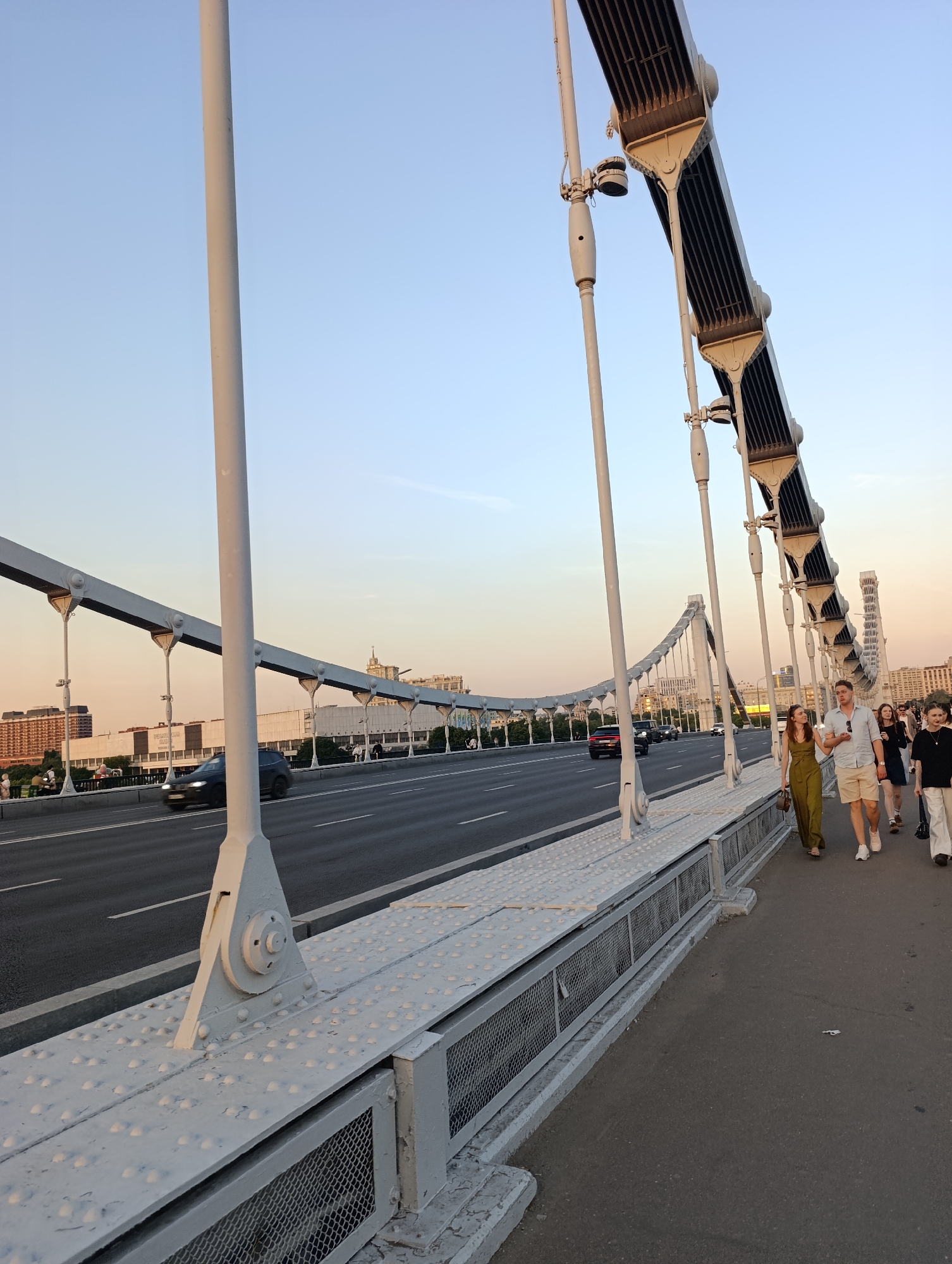 This image captures a wide-angle view of the Krymsky Bridge (Crimean Bridge) in Moscow, Russia, during what appears to be the golden hour, evidenced by the warm hues in the sky. The perspective is from the pedestrian walkway on the right side of the bridge, looking towards the south-east. In the immediate foreground, the robust, light grey steel structure of the bridge is prominent. This includes a massive vertical mast or suspension rod painted light grey, securely bolted to a triangular base plate on the pedestrian deck. The deck itself is composed of riveted or bolted metal plates with a studded surface pattern, adjacent to a mesh-paneled barrier separating it from the roadway. To the right, on the asphalt pedestrian walkway, several people are visible. In the foreground, a woman in an olive-green jumpsuit with a brown handbag walks alongside a man in a light blue short-sleeved shirt and beige shorts, both wearing light-colored shoes. They appear to be conversing and walking leisurely. Behind them, further down the walkway, a woman in a dark top and light pants, and another individual in a dark dress, are also walking. To the left of the pedestrian walkway, separated by a low concrete barrier, is the asphalt roadway. Multiple lanes are visible, carrying vehicular traffic. A dark-colored SUV, possibly a BMW X model, is prominent in the mid-ground, traveling away from the viewer, exhibiting slight motion blur. Other vehicles are present further down the road. The bridge's distinctive suspension system is visible, featuring a series of large, light grey, box-girder-like segments from which vertical tension rods extend downwards to support the bridge deck. These segments are connected by intricate fastenings. Another set of suspension cables or rods arc upwards and outwards from the main deck, forming the characteristic silhouette of the Krymsky Bridge. In the background, a panorama of Moscow's urban landscape unfolds under a sky that transitions from light blue overhead to a warm, soft peach and yellow hue closer to the horizon. Notable buildings include the New Tretyakov Gallery (Central House of Artists), identifiable by the Cyrillic text "ТРЕТЬЯКОВСКАЯ ГАЛЕРЕЯ" on its façade. Other commercial and residential buildings with varying architectural styles and heights are visible across the skyline. A golden dome, likely belonging to the Cathedral of Christ the Savior, is faintly discernible on the distant horizon. The overall scene depicts urban activity on a major infrastructure landmark under pleasant