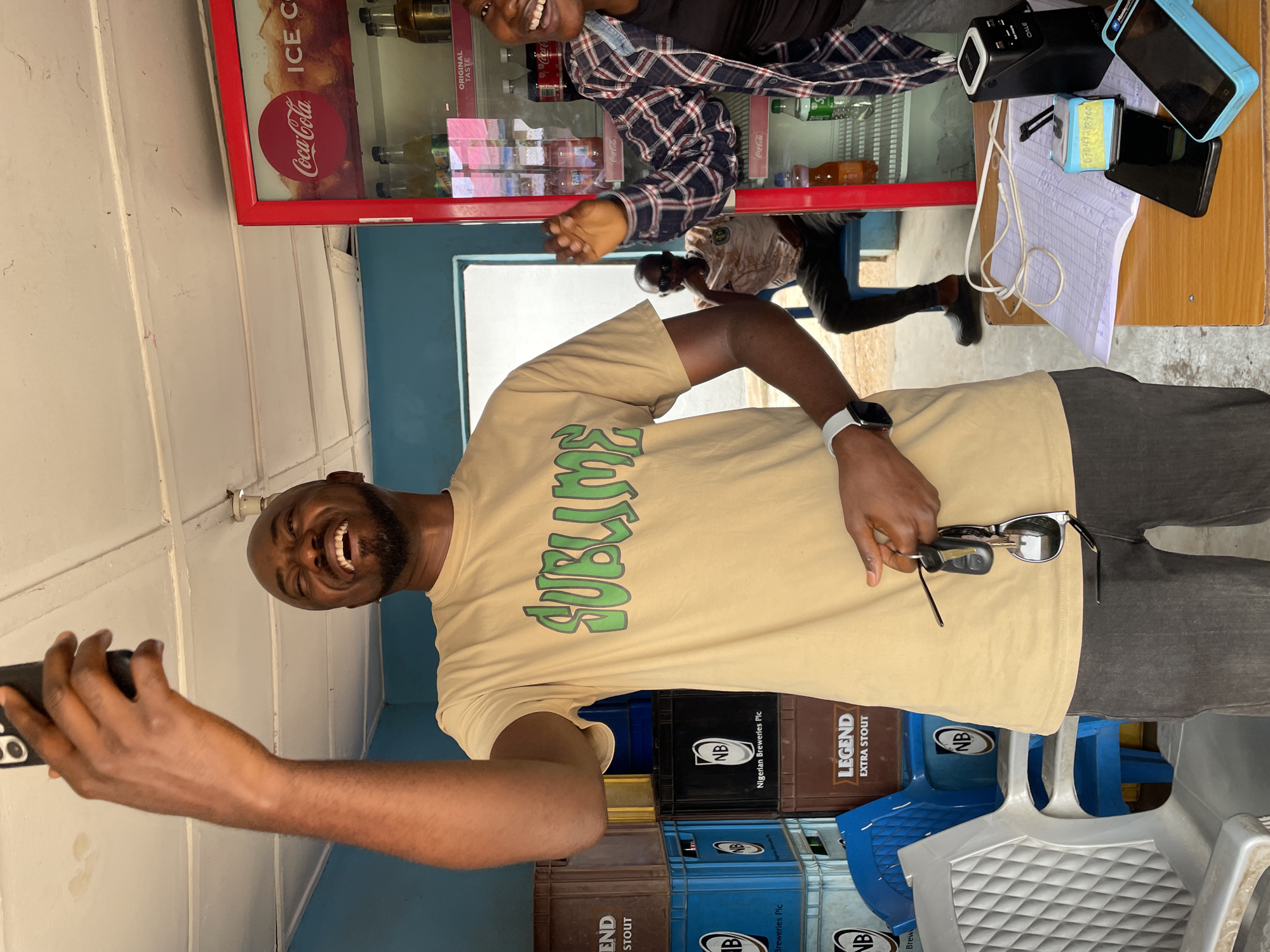 A lively scene unfolds inside what appears to be a shop or convenience store in Jos, Nigeria, during the daytime. The primary subject is a smiling man with a beard, wearing a beige t-shirt with "SUBLIME" printed in green. He is holding a black smartphone, seemingly taking a selfie or video, while also holding sunglasses and keys in his other hand. A white smartwatch is visible on his wrist.
Behind him, another person with a bright smile, wearing a plaid shirt, is partially visible near a red-framed Coca-Cola display fridge. The fridge contains various bottled beverages and features "Coca-Cola," "ICE C," and "ORIGINAL TASTE" text. Further back, another individual is faintly seen standing behind a counter.
The counter, made of wood, holds several items: a blue mobile phone (possibly a Microsoft device), a black mobile phone, a black device labeled "OALE", a white sheet of paper with writing, a white charging cord, and a blue stamp with the number "07049298900".
At the bottom of the frame, numerous crates are stacked, some blue and some brown, containing beverages. Visible text on the crates includes "NB", "Nigerian Breweries Plc", "LEGEND EXTRA STOUT", and "BREW". Plastic chairs are also visible nearby. The overall atmosphere conveyed is cheerful, with both visible subjects smiling broadly. The background features plain light-colored walls and a light blue section, indicating an indoor or semi-enclosed setting.