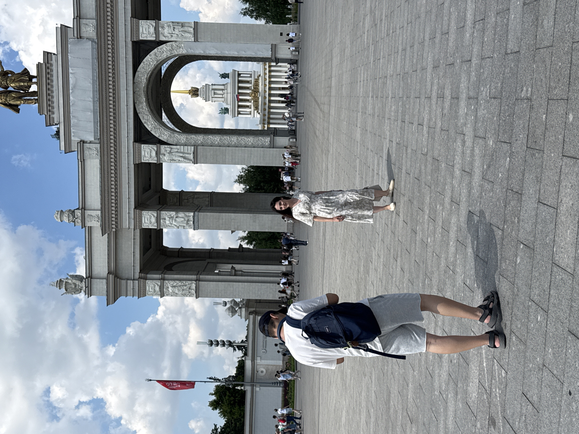 The image shows a wide, paved plaza in front of a large, ornate building with classical architectural elements, including arches and columns. In the foreground, a man wearing a white t-shirt, grey shorts, a dark baseball cap, and a dark backpack is standing and looking down at his phone. To his right, a woman in a light-colored patterned dress and white shoes is standing and looking towards the camera. In the background, through the archway of the building, another prominent building with a golden spire is visible. Several other people are scattered throughout the plaza, walking or standing, and there are trees and a red flag visible in the distance. The sky is blue with white, fluffy clouds. This scene appears to be the VDNKh (Exhibition of Achievements of National Economy) in Moscow, Russia, a large exhibition center known for its Soviet-era architecture.