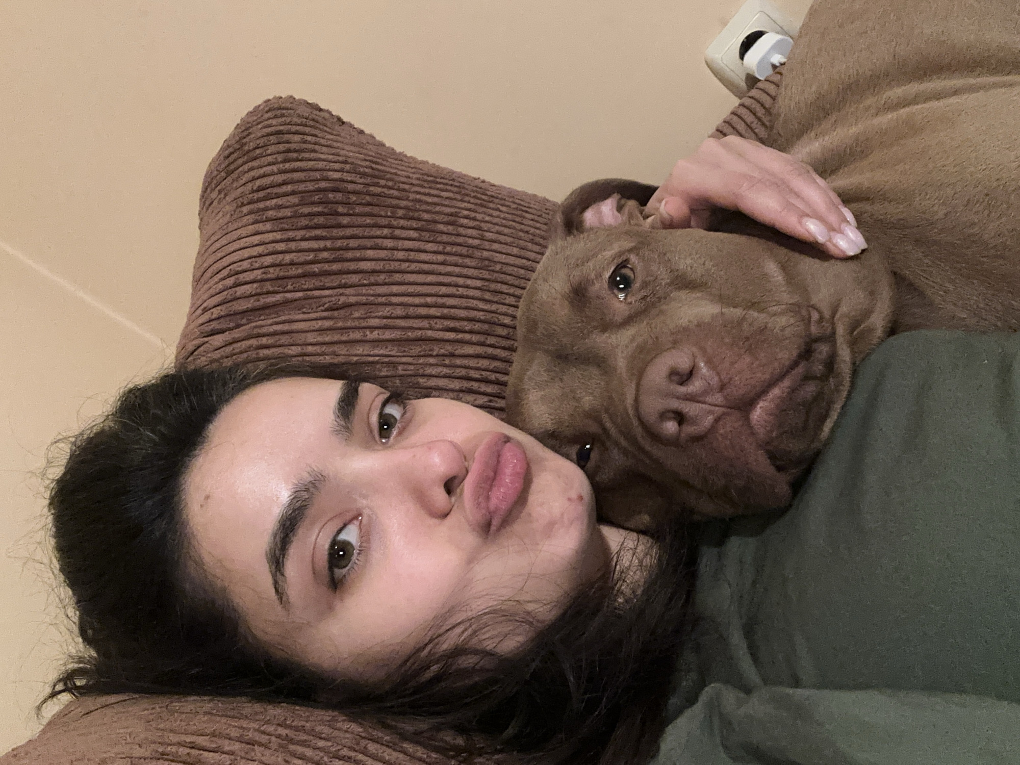 A young woman with dark hair and brown eyes is taking a selfie with a brown pit bull. She is puckering her lips and has her arm around the dog's neck. The dog is resting its head on her chest, and its eyes are looking directly at the camera. They are both on a brown, corduroy-covered sofa. The background is a plain beige wall with a white electrical outlet. The overall setting appears to be a living room. The image is not taken in Malevo, Bulgaria.