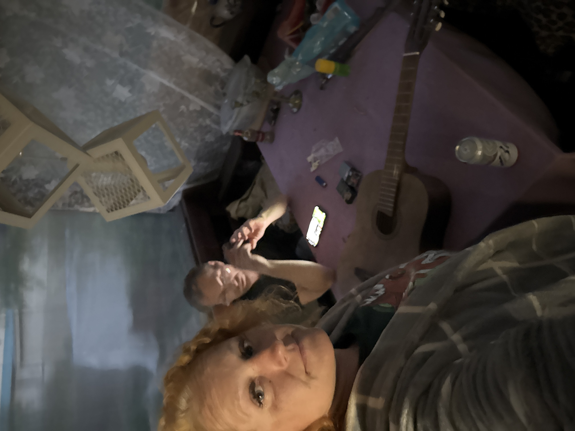 The image depicts an indoor scene with two individuals and various objects on a table. In the foreground, a woman with red, curly hair is visible, facing the camera. She is wearing a grey plaid blanket draped over her shoulders. Behind her, a man with short brown hair is seated, looking towards the camera. He is wearing a dark t-shirt.
The table in the center of the image is covered with a purple tablecloth. On the table, there are several items, including a guitar, a can of soda, a cell phone, lighters, a bottle of alcohol, and a glass bowl. The background shows a wall with a mural or painted scenery, possibly depicting a waterfall or natural landscape. There are also decorative elements on the wall, including what appear to be cube-shaped decorations and sheer curtains with a star pattern. The overall environment appears to be a dimly lit room, possibly a living space or a gathering area. The location is identified as Chaikovsky, Russia.