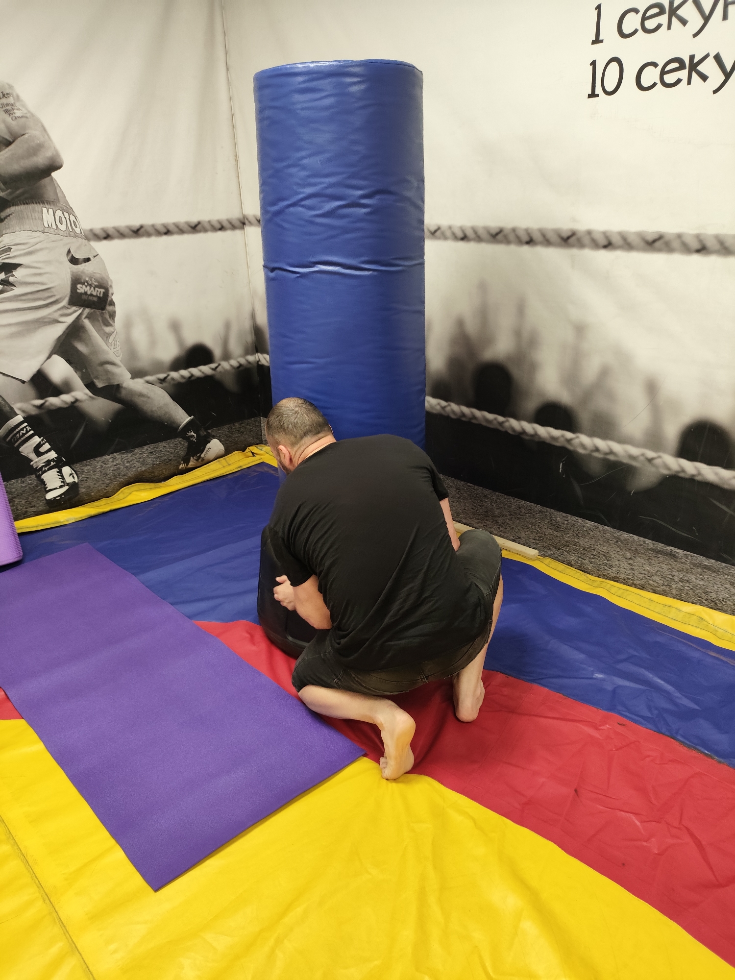 A man wearing a black t-shirt and black shorts is kneeling on a colorful mat in what appears to be a gym or training facility. He is facing away from the camera and is in the process of interacting with a large, black, cylindrical object, possibly a weighted bag or a tire. The mat is sectioned into red, blue, and yellow colors. To the left of the man, a purple mat is visible. In the background, a large mural depicts a boxing ring and boxers, with some text in Russian on the wall above. The overall environment suggests a martial arts or fitness training space.