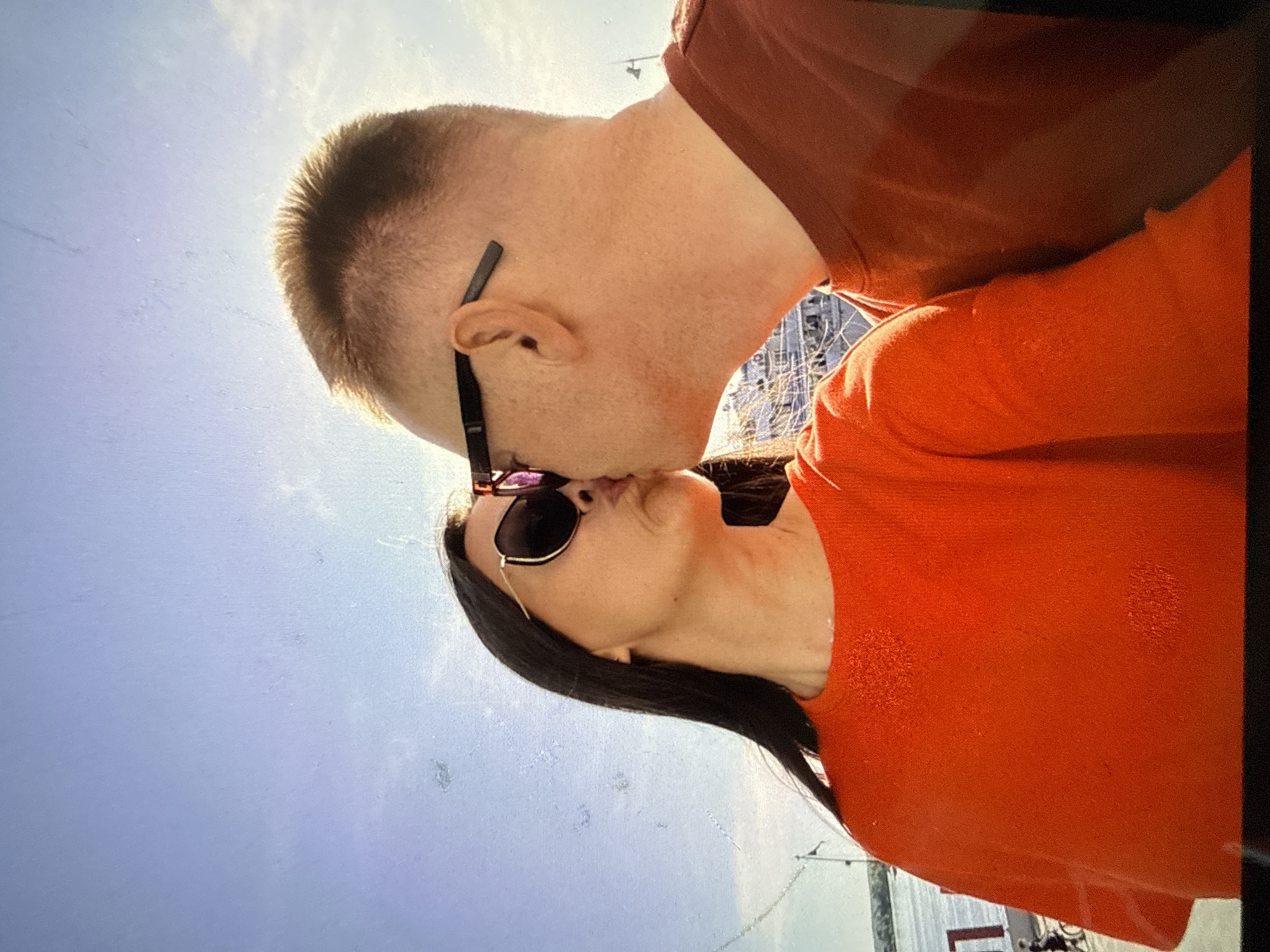 An outdoor, waist-up, medium shot shows a couple kissing on a sunny day. The person on the left is a woman with long dark hair wearing large, dark sunglasses and an orange crewneck sweater. She is leaning towards the person on the right. The person on the right is a man with short, light brown hair, wearing dark sunglasses and a rust-colored t-shirt. His head is turned towards the woman, and their lips are touching. They are in front of a bright blue sky with some distant clouds. In the background, there are hints of a body of water and what appears to be a ship or large structure, possibly a pier or riverboat, with some railings and masts visible. The environment suggests a public outdoor space in Chaikovsky, Russia, likely near the Kama River, given the presence of water and the type of background structures often found along riverbanks or ports in the city.