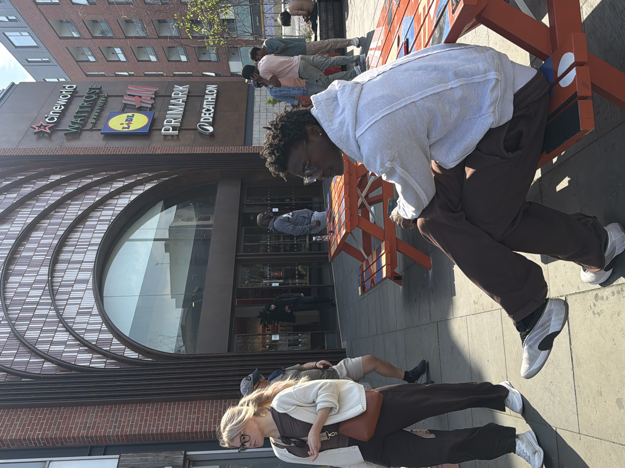 This media file captures a sunny daytime street scene in London, United Kingdom, showing a modern commercial building and several people. In the immediate foreground, a person with dark, curly hair and glasses, wearing a light grey hoodie and dark brown sweatpants, is leaning over an orange wooden public bench or structure. This individual is looking downwards, possibly at a phone. The bench features white and blue painted accents. Strong shadows are cast on the light-colored paving stones, indicating bright sunlight.
Behind the foreground subject, several other individuals are visible. To the right, another person is bending over the orange structure, shirtless. Further back, a small group of people are standing or walking; one is wearing a light pink shirt and khaki pants, and another in a light blue shirt. Towards the bottom left, partially cropped, a blonde woman in a white cardigan, dark top, and dark pants, carrying a brown shoulder bag and wearing glasses, is seen. Next to her is a man in a dark baseball cap. Near the building's main entrance, a person in a grey hoodie is also visible.
The background features a multi-story brick building with a large, arched glass entrance on the ground floor. Above this entrance, prominent signs for various retailers are displayed vertically: "cineworld" (with a red star logo), "WAITROSE & PARTNERS", "LIDL" (in its distinctive blue and yellow square logo), "PRIMARK", and "DECATHLON". The upper floors of the building have multiple rectangular windows. The overall impression is a bustling urban commercial area on a clear day.