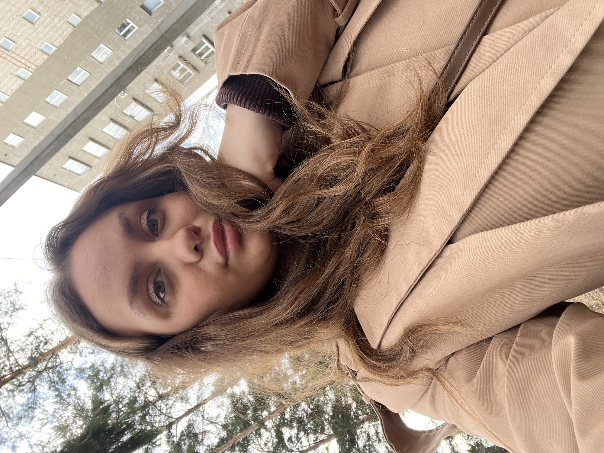 A woman with light brown hair is wearing a tan trench coat and a brown sweater. The background includes a tall, concrete building with numerous windows and bare trees. The location is likely Chaikovsky, Russia.