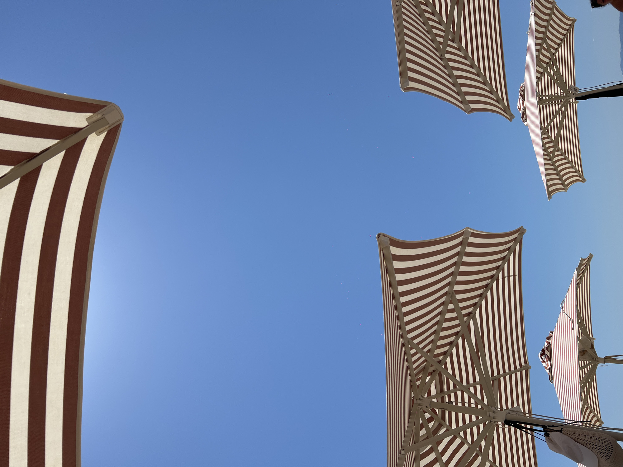 In Troulos, Greece, under a clear blue sky, several striped beach umbrellas are angled upwards. One umbrella is partially visible at the top of the frame, with brown and white stripes. In the lower half of the image, two more umbrellas are open, their striped canopies and supporting structures visible against the sky. To the left, another umbrella is seen from below, with a woven bag and a white hat hanging from its pole. A fourth umbrella is positioned in the lower right, partially obscured by other elements. The scene is set outdoors, suggesting a beach or a similar recreational area on a sunny day.