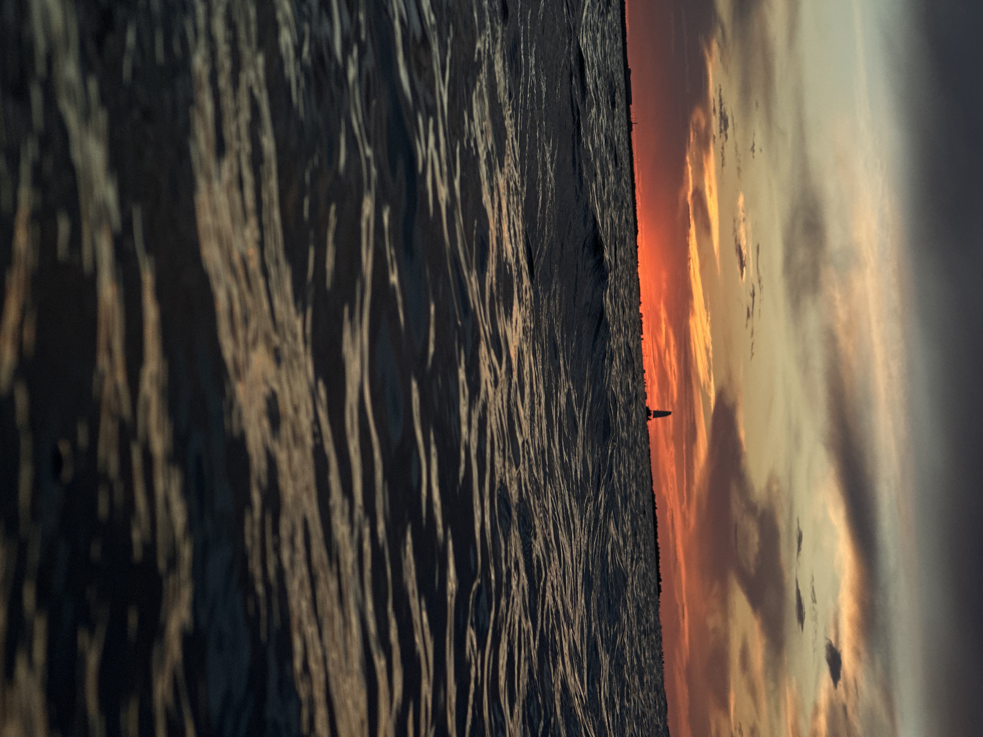 The image captures a sunset over a body of water, with a silhouette of a sailboat visible on the horizon. The sky is filled with dramatic clouds in shades of orange, red, and gray. The water in the foreground is dark and choppy, reflecting the colors of the sky. The scene is tranquil and serene, evoking a sense of peace and solitude. The location is not specified in the image, and therefore cannot be determined.