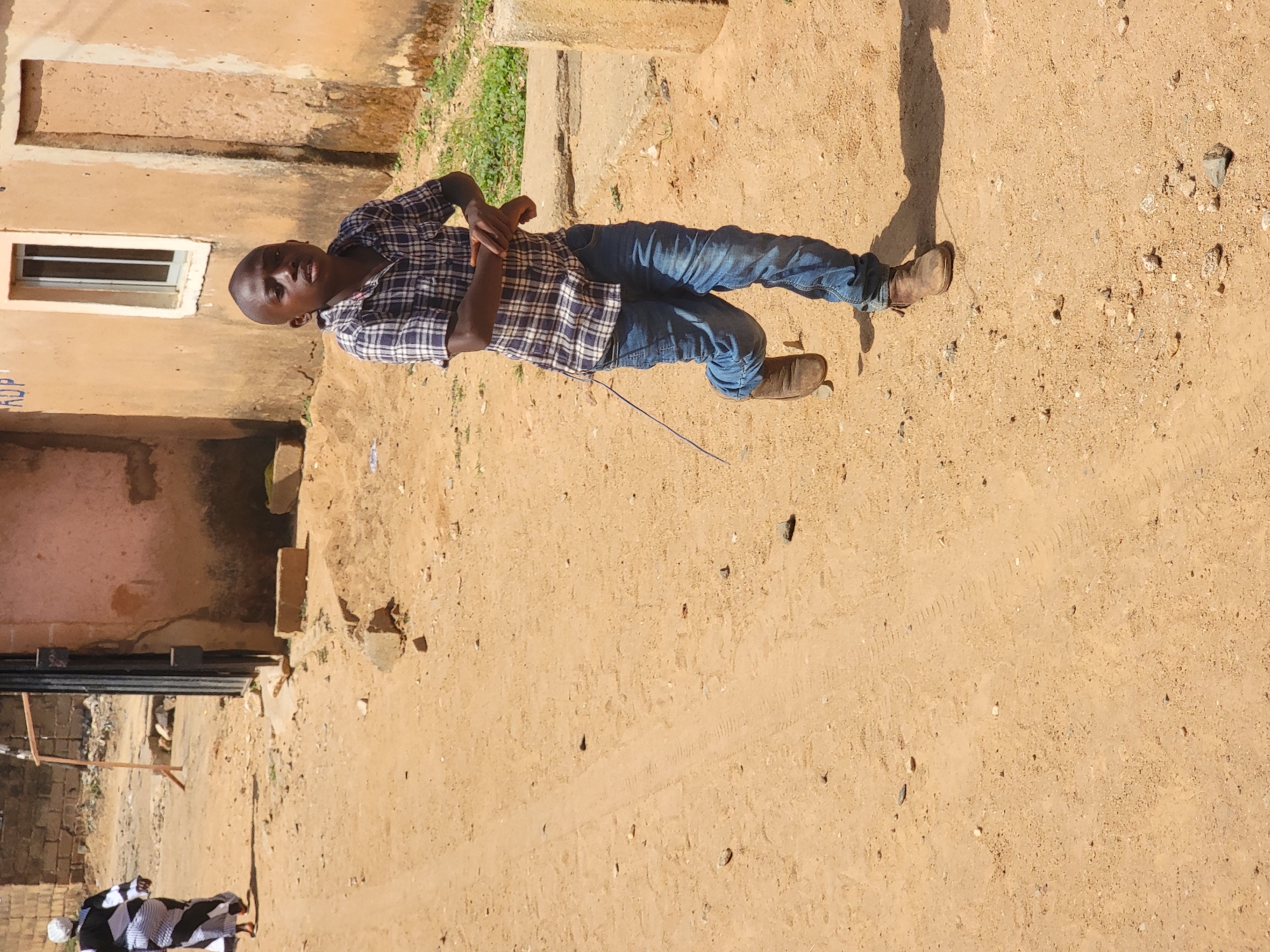 A man stands on a dirt road in what appears to be a residential area of Jos, Nigeria. He is wearing a plaid shirt, jeans, and brown shoes. The man is gesturing with his hands and looking toward the camera. Behind him is a building with a window and a fenced area. The dirt road shows signs of tire tracks. Another person can be seen in the lower left corner of the image wearing what appears to be a black and white patterned dress. The lighting suggests a sunny day.