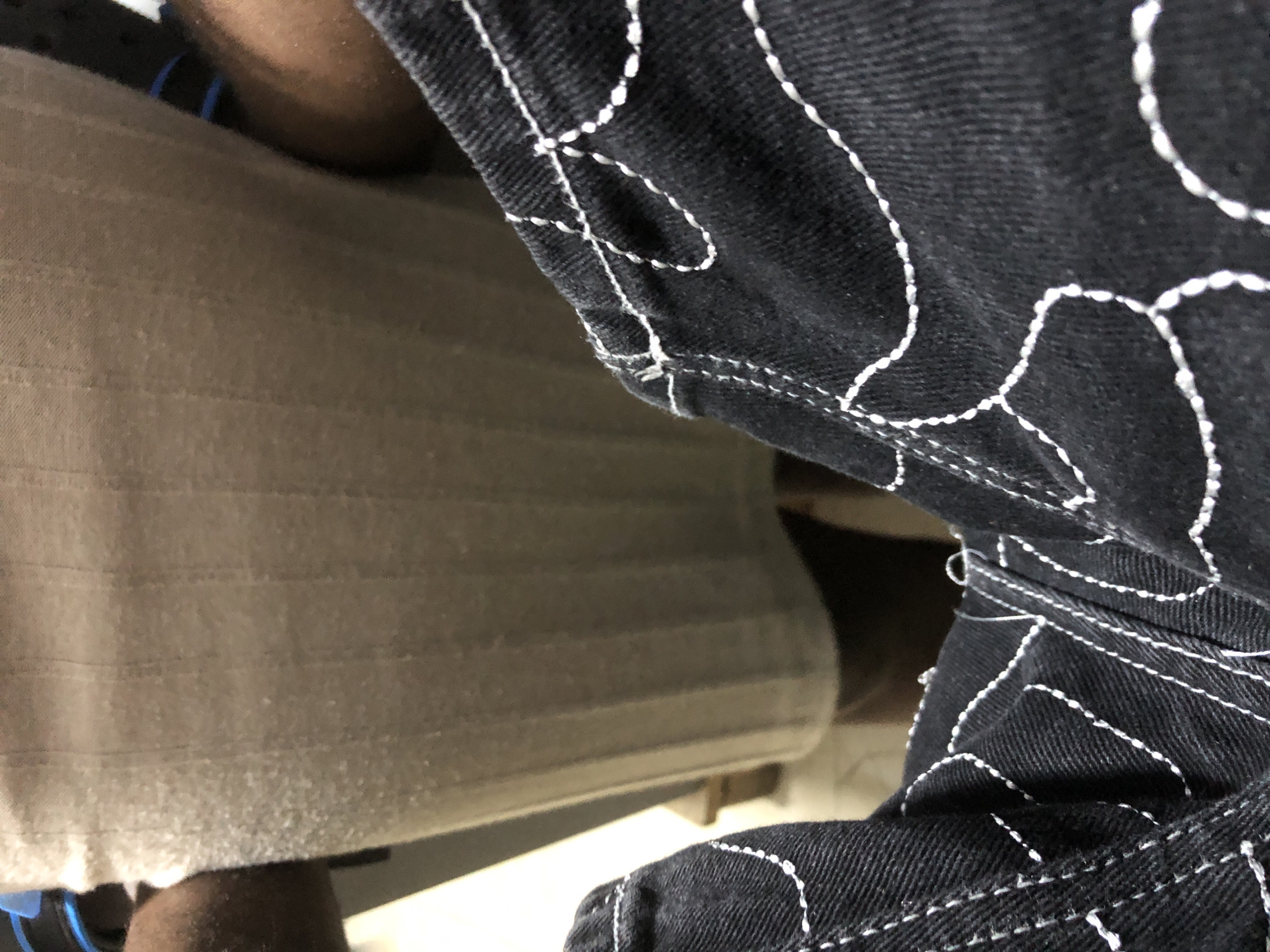 The image shows a close-up view of dark denim jeans with white stitching. The stitching forms an abstract, wavy pattern across the fabric. In the background, a person's legs are visible, wearing a tan-colored garment. The environment appears to be indoors, possibly in a building. The lighting is dim, casting shadows. There is no indication of the specific location within Ado, Nigeria, or any context to determine the action or interaction taking place.