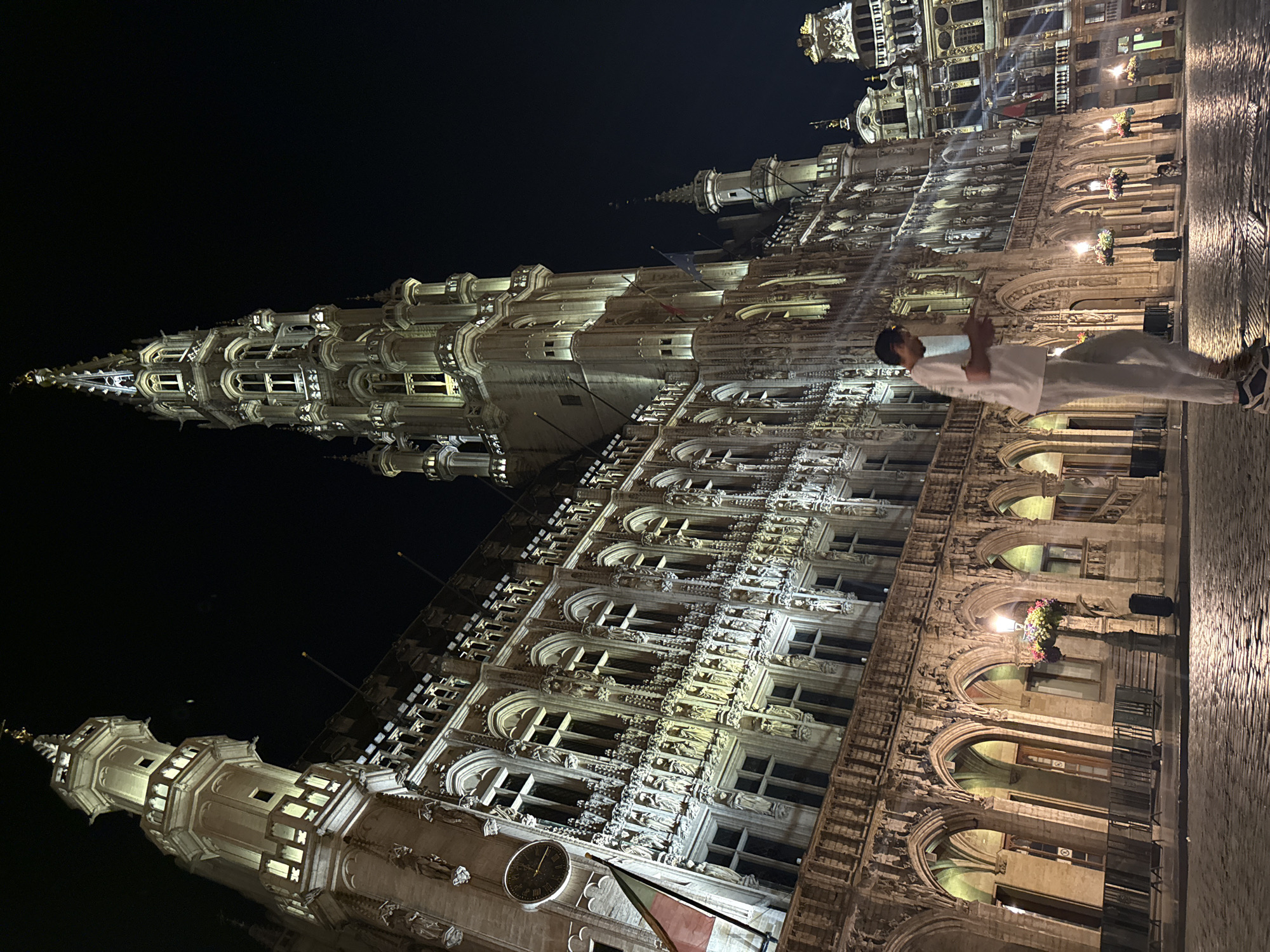 The image captures a nighttime view of the Grand Place in Brussels, Belgium. The focal point is the ornate facade of the Town Hall, illuminated by warm lighting that highlights its intricate Gothic architecture. A tall, spired tower dominates the right side of the frame, also lit from below. In the foreground, a cobblestone plaza reflects the ambient light, with a few lampposts adorned with flower baskets. A young person, dressed in light-colored athletic wear, is walking across the plaza, looking down at their phone. Other figures are faintly visible in the background, also dispersed across the plaza. The sky is dark, indicating it is nighttime.