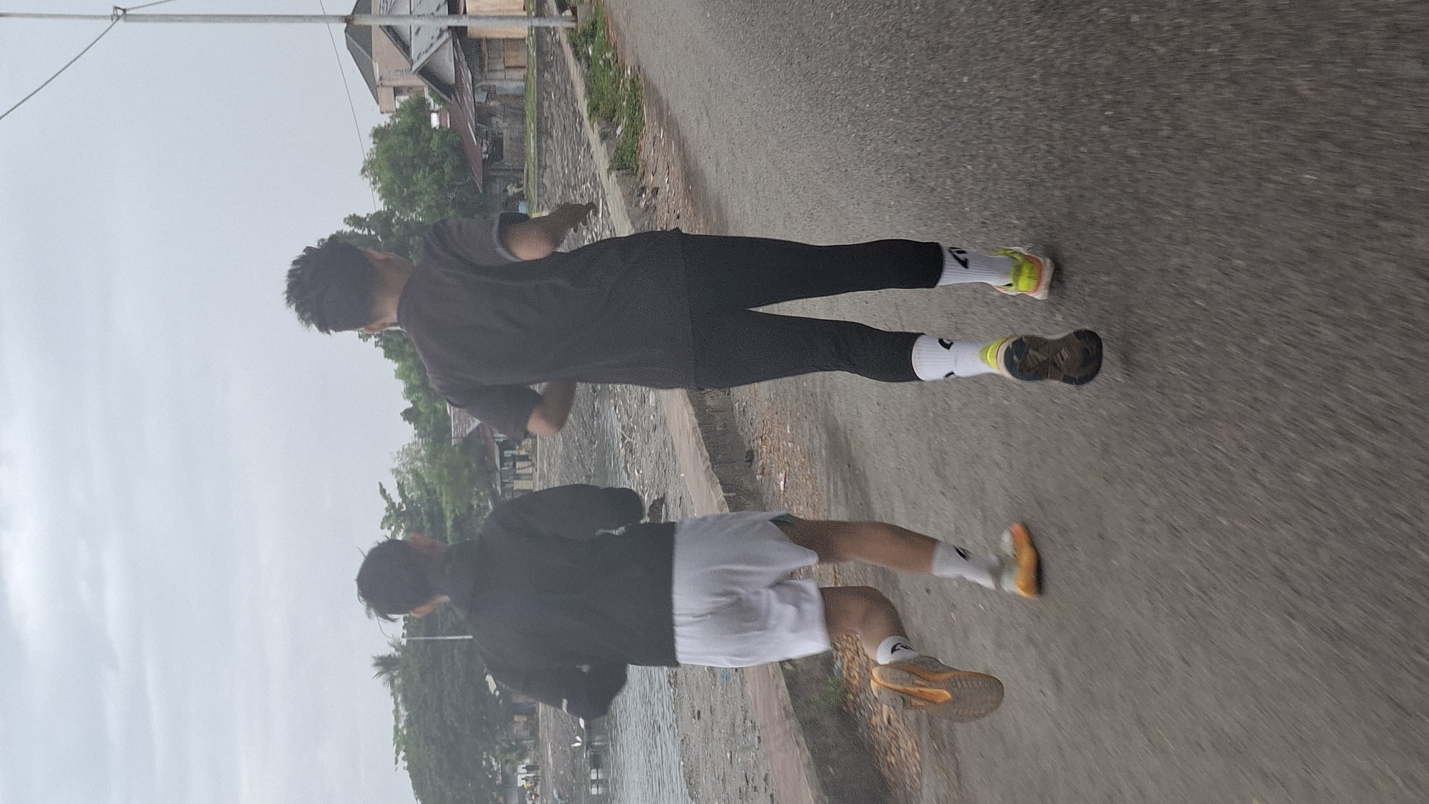Two individuals, viewed from behind, are jogging on a paved road. The person on the left wears a black jacket over white shorts and bright orange running shoes with white socks. The person on the right is dressed in a black t-shirt, black leggings, and yellow and black running shoes with white socks. The road appears to be adjacent to a body of water on the left, with a concrete barrier separating the road from the water's edge. In the background, there are trees, scattered buildings with tin roofs, and a cloudy sky. This scene is consistent with a location in Lhokseumawe, Indonesia, likely near the coast or a riverbank. The image captures a moment of physical activity in an urban or semi-urban coastal environment.