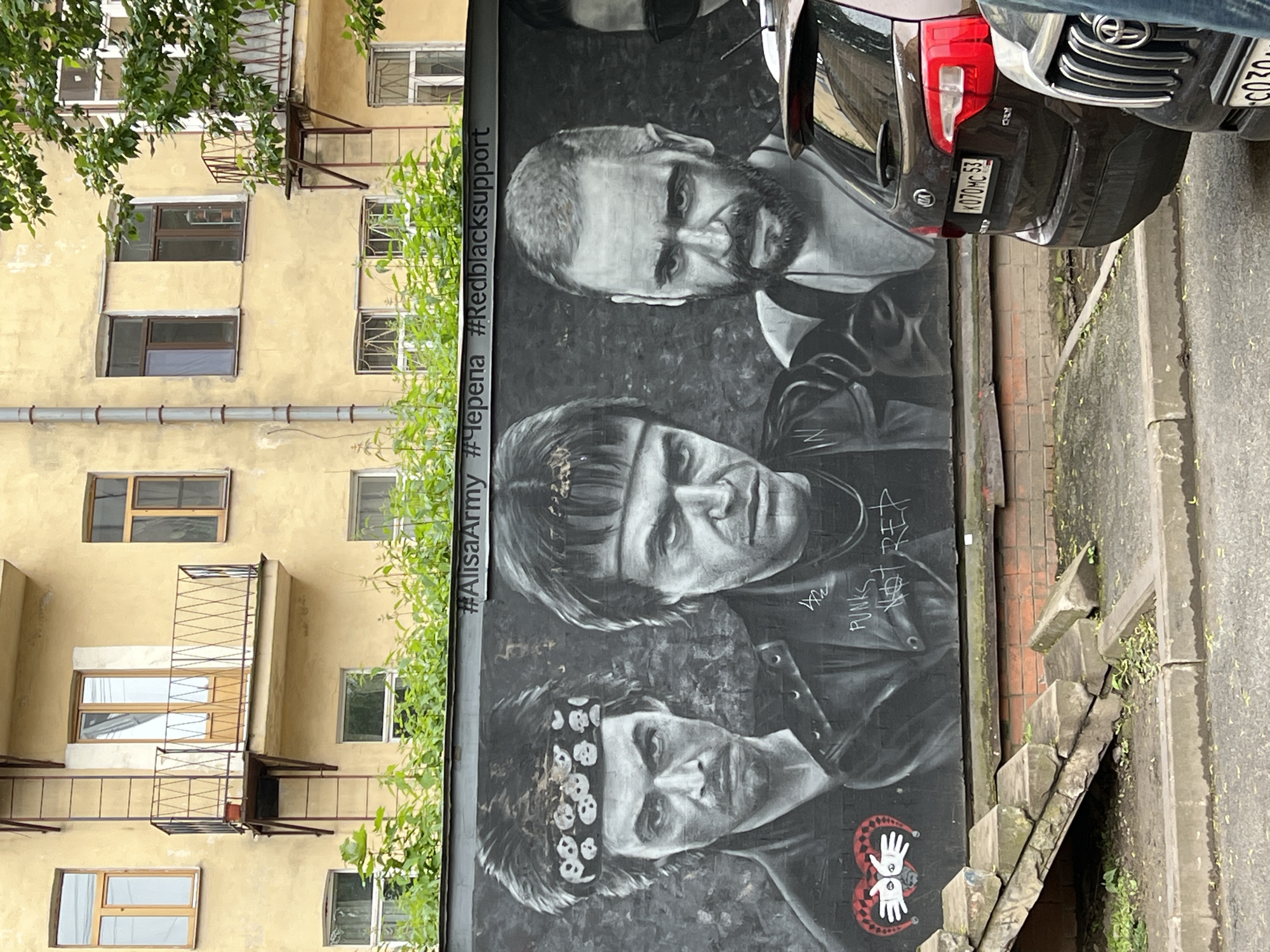 The image shows a black and white mural on a wall next to a building in Saint Petersburg, Russia. The mural features three male figures in a portrait-like arrangement. The man at the top has a beard and a somewhat intense gaze. The man in the middle has bangs and is wearing a leather jacket. The man at the bottom has a bandana with skull motifs. The mural includes text "#AlisaArmy #Черепа #Redblacksupport" and the phrase "DAÑ PUNKS NOT DEAD" written in a graffiti style. The mural appears to be near street level, with visible steps leading down to an unseen area next to it. A black car is parked adjacent to the mural, partially obscuring the top right corner of the image. The building next to the mural has yellow walls with windows and a fire escape. Trees are visible on the left side of the image. The setting appears urban.