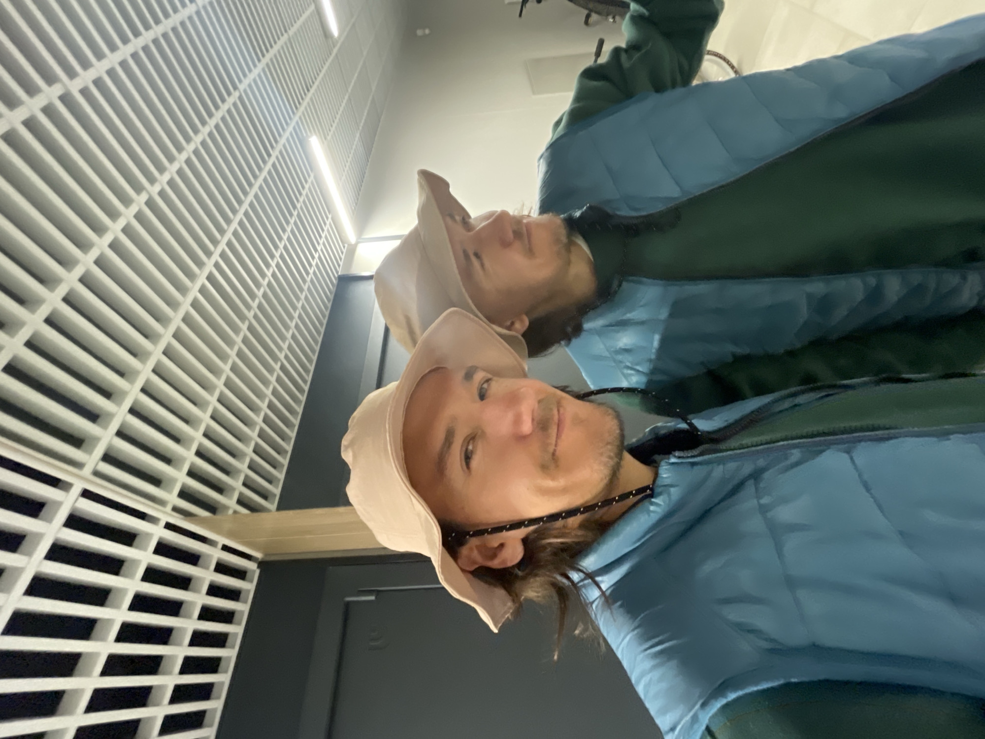A medium shot of a man wearing a tan bucket hat, a blue puffy vest, and a green undershirt standing indoors. The man has tan skin, brown hair, and a mustache. He has a rope attached to his hat and eyeglasses. The image appears to be a selfie. In the background is a person with a tan bucket hat and blue puffy vest, with what appears to be a bicycle above them. Behind the man, on the left, are rows of white, square panels with square holes cut out of them. The walls appear to be gray.