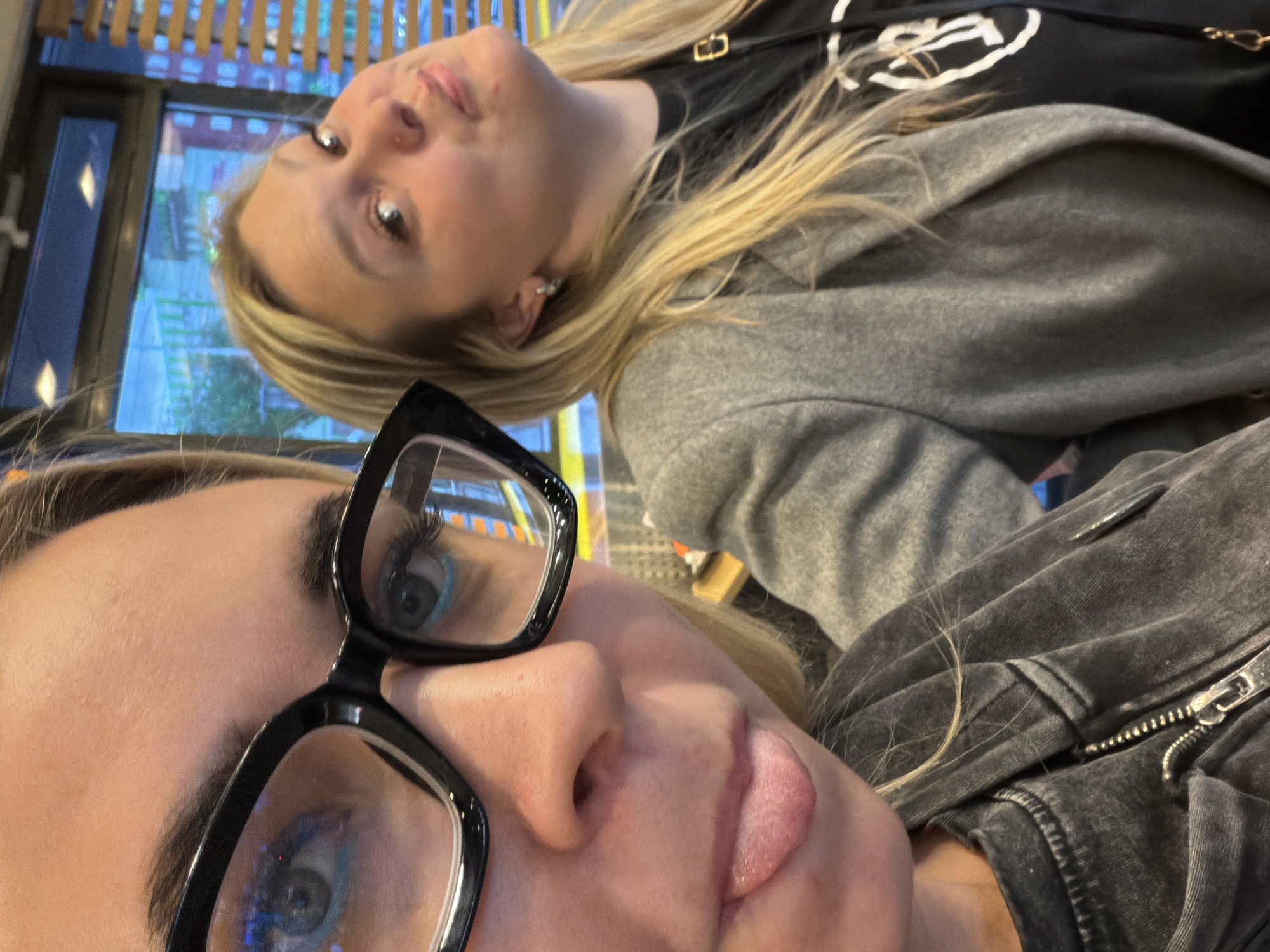 The image shows two light-skinned women posing closely for a selfie. The woman in the foreground wears black glasses and has her tongue slightly sticking out. She is wearing a gray jacket with a zipper. The other woman is wearing a gray sweatshirt over a black t-shirt and has blonde hair and an earring in her right ear. The background appears to show a window with light coming through and a cityscape. There is no information available that indicates the location in Izhevsk, Russia.