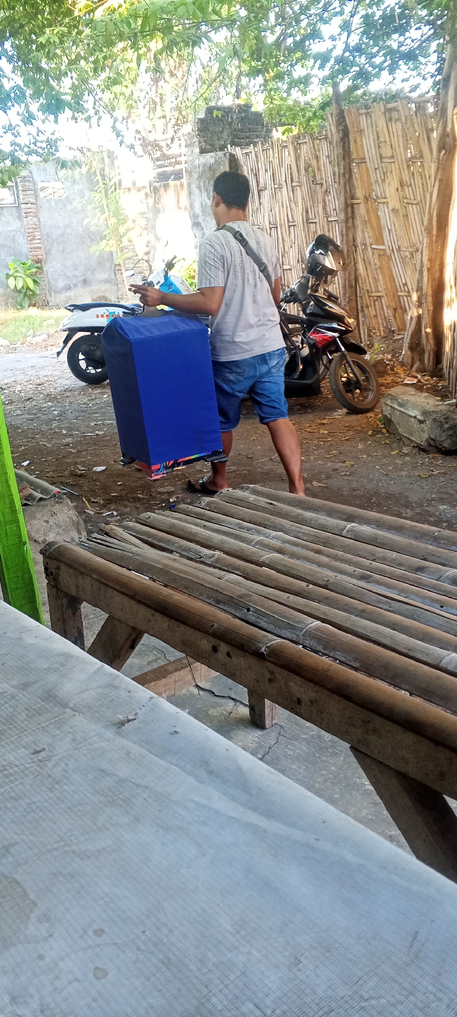 The image depicts an outdoor scene in Lumajang, Indonesia, likely a residential area or a yard. A man wearing a light-colored t-shirt and denim shorts is carrying a large, blue, rectangular cage. The cage appears to be made of fabric stretched over a frame and has a colorful patterned base visible at the bottom. In the background, a motorcycle is parked, with a helmet on its seat. There is a bamboo fence and some structures that suggest buildings, possibly made of concrete and brick. A rustic wooden bench with bamboo slats is in the foreground. The ground is dirt and gravel, with some vegetation visible. The lighting suggests it is daytime, with sunlight filtering through the trees.