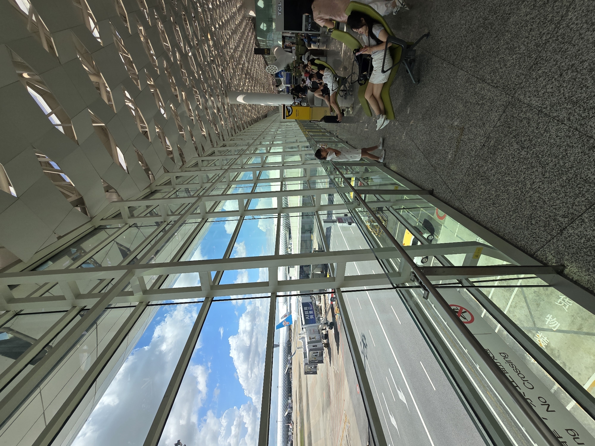 This image depicts the interior of an airport terminal in Bao'an District, China, with a large glass window overlooking the tarmac. Through the window, an airplane is visible parked at a gate, connected to a jet bridge. The terminal interior features a modern architectural design with a high ceiling and extensive use of glass and white structural elements. A row of lounge chairs is arranged along the window, occupied by several people, some of whom are seated and looking at their phones. A young girl stands near the window, looking out. The floor is a speckled grey tile, and the overall atmosphere appears to be one of waiting or transit. The lighting suggests it is daytime.