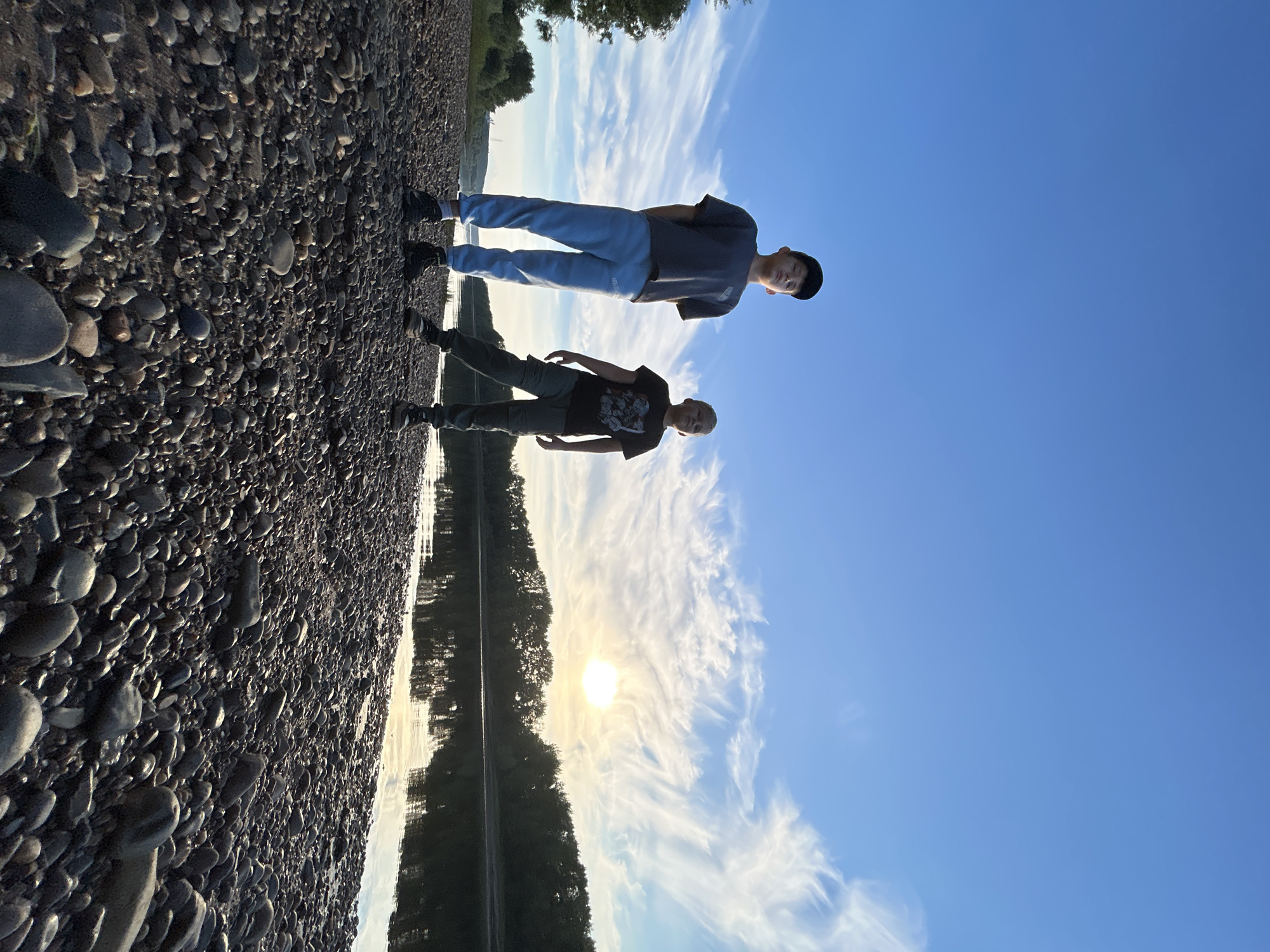 Two young boys stand on a rocky riverbank in Chaikovsky, Russia. The boy on the left wears a dark grey t-shirt, light blue sweatpants, and a black baseball cap. The boy on the right wears a dark t-shirt with a graphic print and dark green cargo pants. They are positioned in the foreground, facing the camera, with their legs slightly spread apart. Behind them, a wide, calm river reflects the sky and the treeline on the opposite bank. The sun is low in the sky, casting a warm glow and creating wispy clouds. The overall scene depicts a serene natural setting during what appears to be sunset or sunrise.