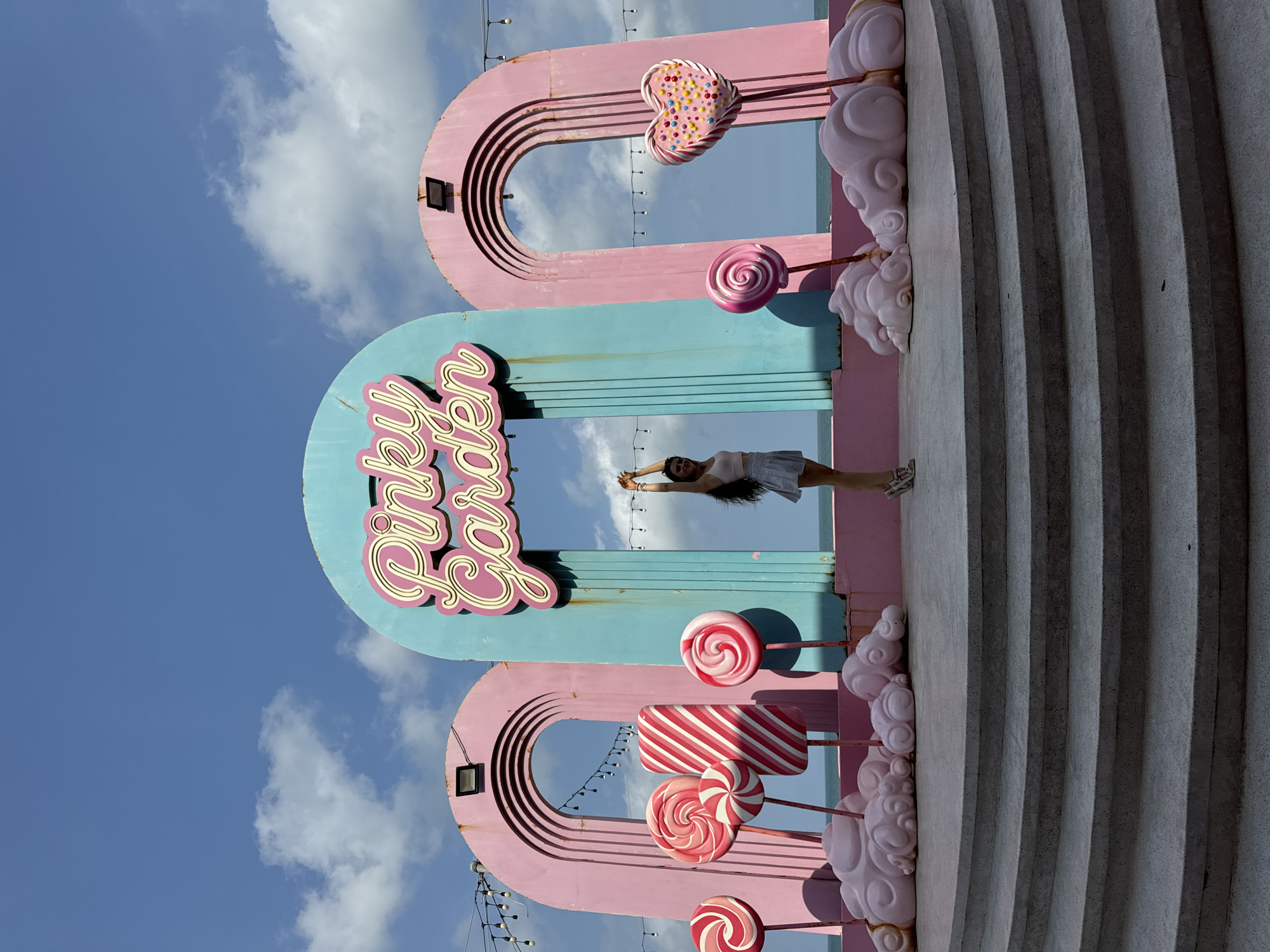 In Tien Thanh Ward, Vietnam, a woman stands on a tiered stone staircase in front of a vibrant, candy-themed display. The display features large, stylized pink and turquoise arches with the neon sign "Pinky Garden" prominently displayed. Flanking the central arch are two pink arches. Scattered throughout the scene are oversized, colorful candy props, including lollipops in red and white, a large striped candy, and a heart-shaped candy adorned with colorful sprinkles. Pink, cloud-like sculptures form the base of the candy props. The woman, wearing a light-colored top and a skirt, has her arms raised above her head, with her back to the camera. The background reveals a bright blue sky with scattered white clouds and a hint of the ocean beyond the display. The overall scene suggests a whimsical and playful outdoor setting.