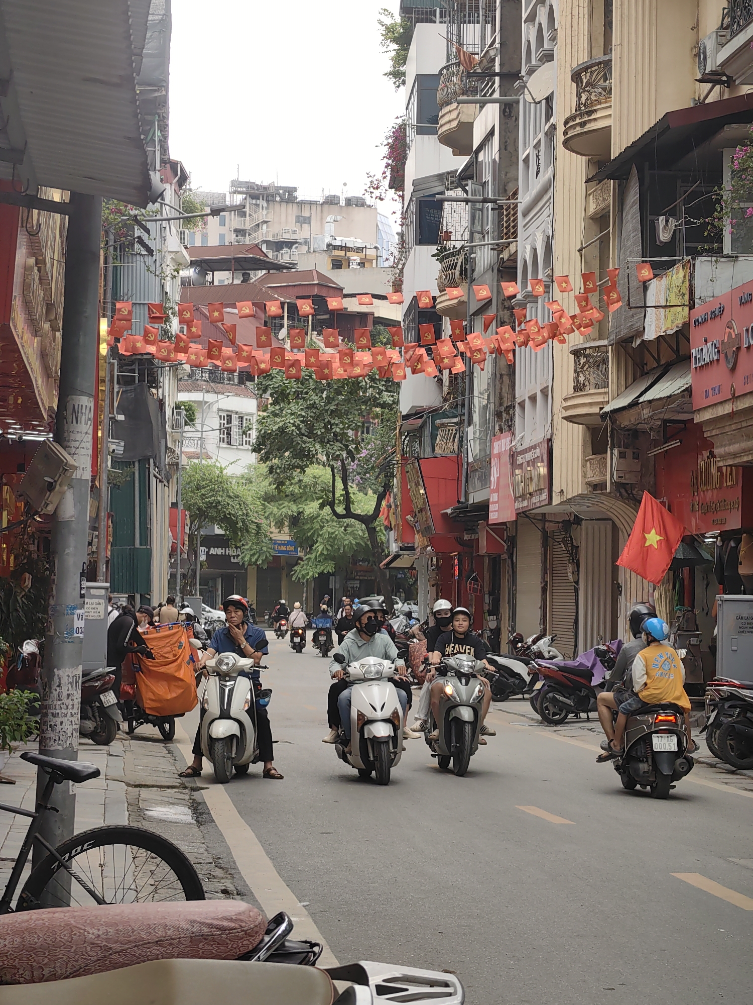 The image depicts a street scene in Hà Nội, Vietnam, characterized by dense urban development and active motorcycle traffic. Overhead, strings of small red Vietnamese flags adorned with a yellow star are strung across the narrow street. The architecture is a mix of older, narrow buildings with balconies and more modern structures. The street is bustling with people riding scooters and motorcycles. Sidewalks are lined with parked motorcycles, bicycles, and various utility poles and street signs. The lighting suggests an overcast day. The overall atmosphere is one of everyday urban life in Vietnam.