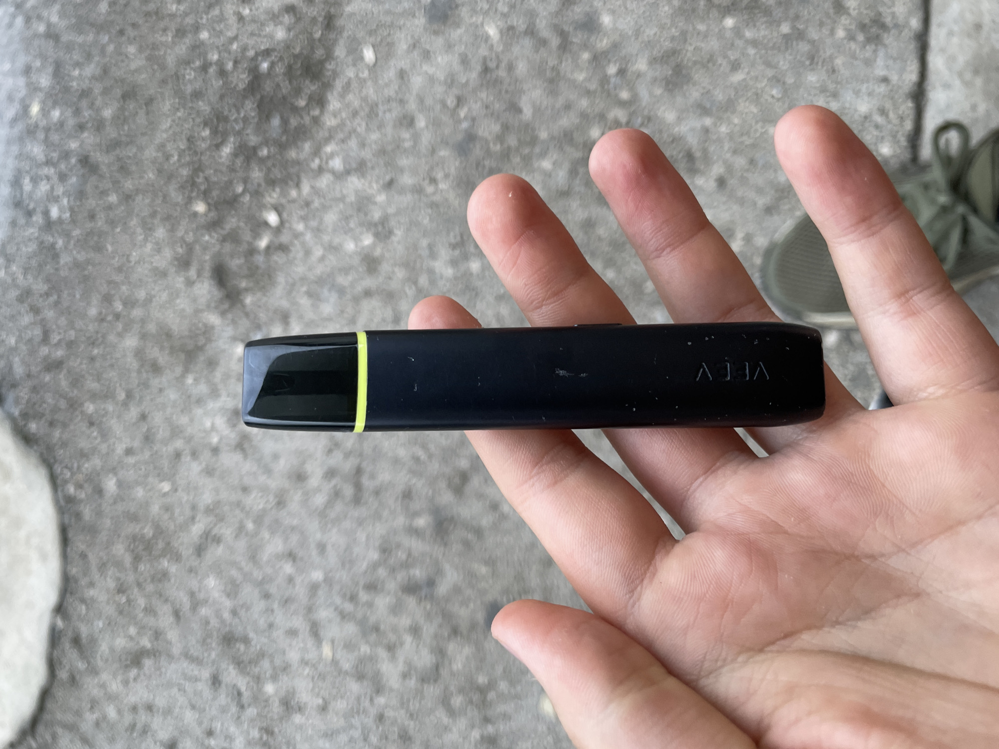 A human hand is prominently featured in the foreground, holding a black electronic device horizontally. The hand has light skin and visible finger creases. The device is rectangular, sleek, and black, with a distinct thin, bright neon yellow stripe on its left end. The brand name "VEEV" is visible in white or light grey lettering on the right side of the device, appearing upside down from the viewer's perspective.
The background consists of a textured, grey surface, likely concrete or pavement, indicating an outdoor setting. Hints of a grey or olive-green shoe are visible in the upper right corner. The lighting suggests it is daytime, with bright and even illumination. No specific activity beyond holding the device is discernible. There are no strong emotional cues.
The image appears to be a close-up shot focusing on the device and the hand. The location provided is Troyan, Bulgaria.