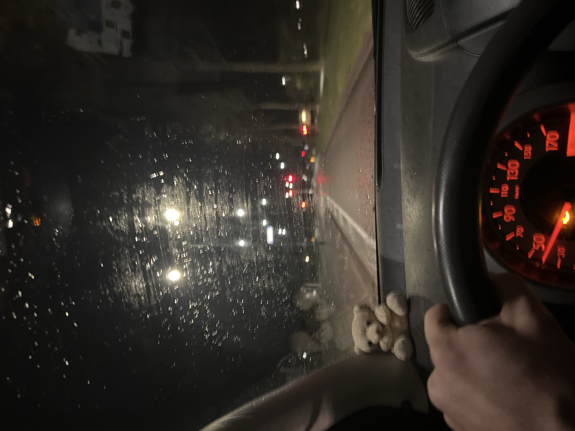 The image displays an interior perspective from a vehicle at night in Geldrop, Netherlands, during rainfall. A human left hand, with a light skin tone, grips the top of a dark, textured steering wheel, partially obscuring the instrument cluster. The speedometer, illuminated in red, shows the needle resting near 0 km/h, with an orange indicator light active at the bottom. A small, light-colored plush teddy bear with a pink nose sits on the dark, slightly dusty dashboard to the left of the driver's perspective.
The windshield is covered in numerous small water droplets, distorting the view ahead. Through the wet glass, a dark, wet asphalt road is visible, reflecting multiple bright streetlights. Several red traffic lights are illuminated in the distance, suggesting the vehicle is stopped at an intersection. White lane markings are discernible on the left side of the road. To the right, a narrow strip of green grass separates the road from a row of dark tree trunks. Faint, blurry outlines of other vehicles, including a white, box-shaped vehicle, are visible further down the road and to the right, integrated into the dark, artificially lit urban environment.